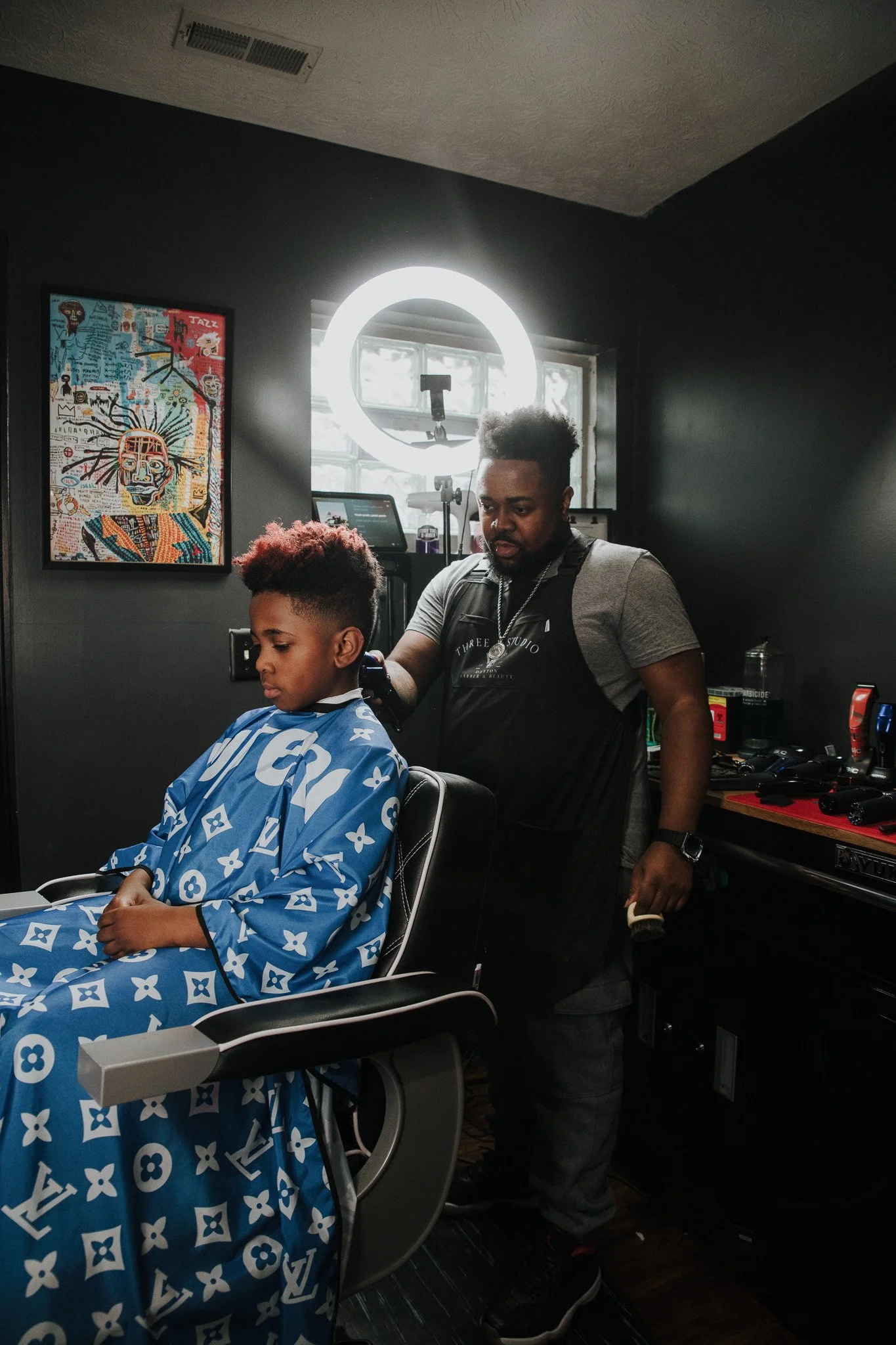 A young boy with dyed hair sitting in a barber chair, wearing a blue Louis Vuitton cape, getting a haircut from a barber in a modern barbershop with dark walls, artwork, and a bright ring light behind them.