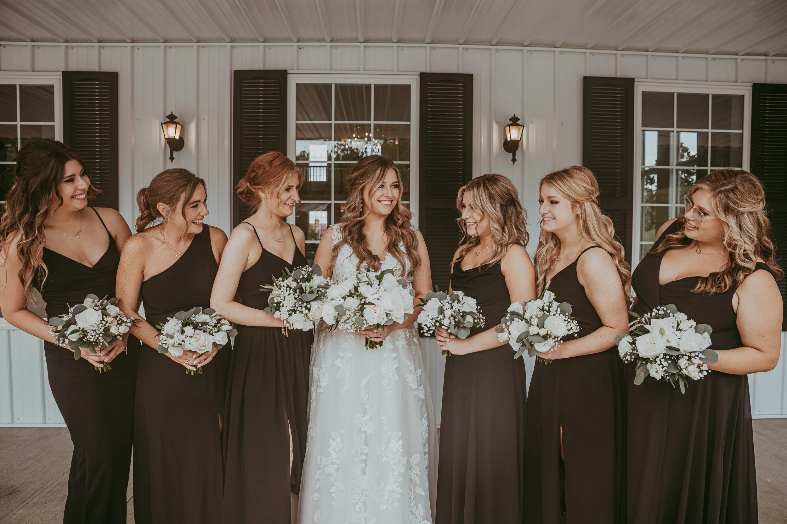 Brunette bride in a white floral gown holding a bouquet, surrounded by seven bridesmaids in black dresses holding matching bouquets, standing in front of a white building with black shutters.