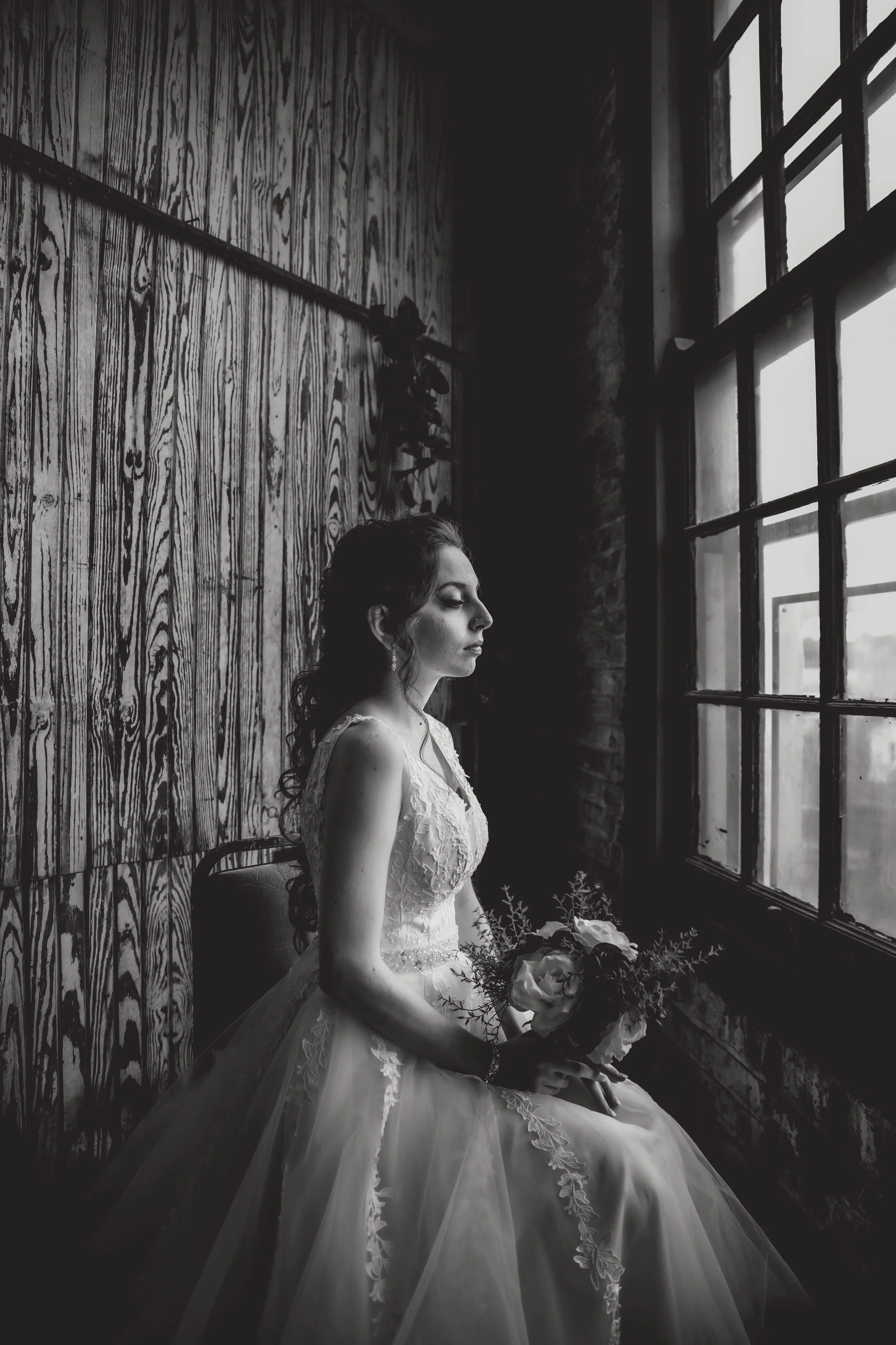 Black and white photo of a woman in a wedding dress sitting by a window, holding a bouquet of flowers, with her eyes closed, in a rustic room with wooden walls.