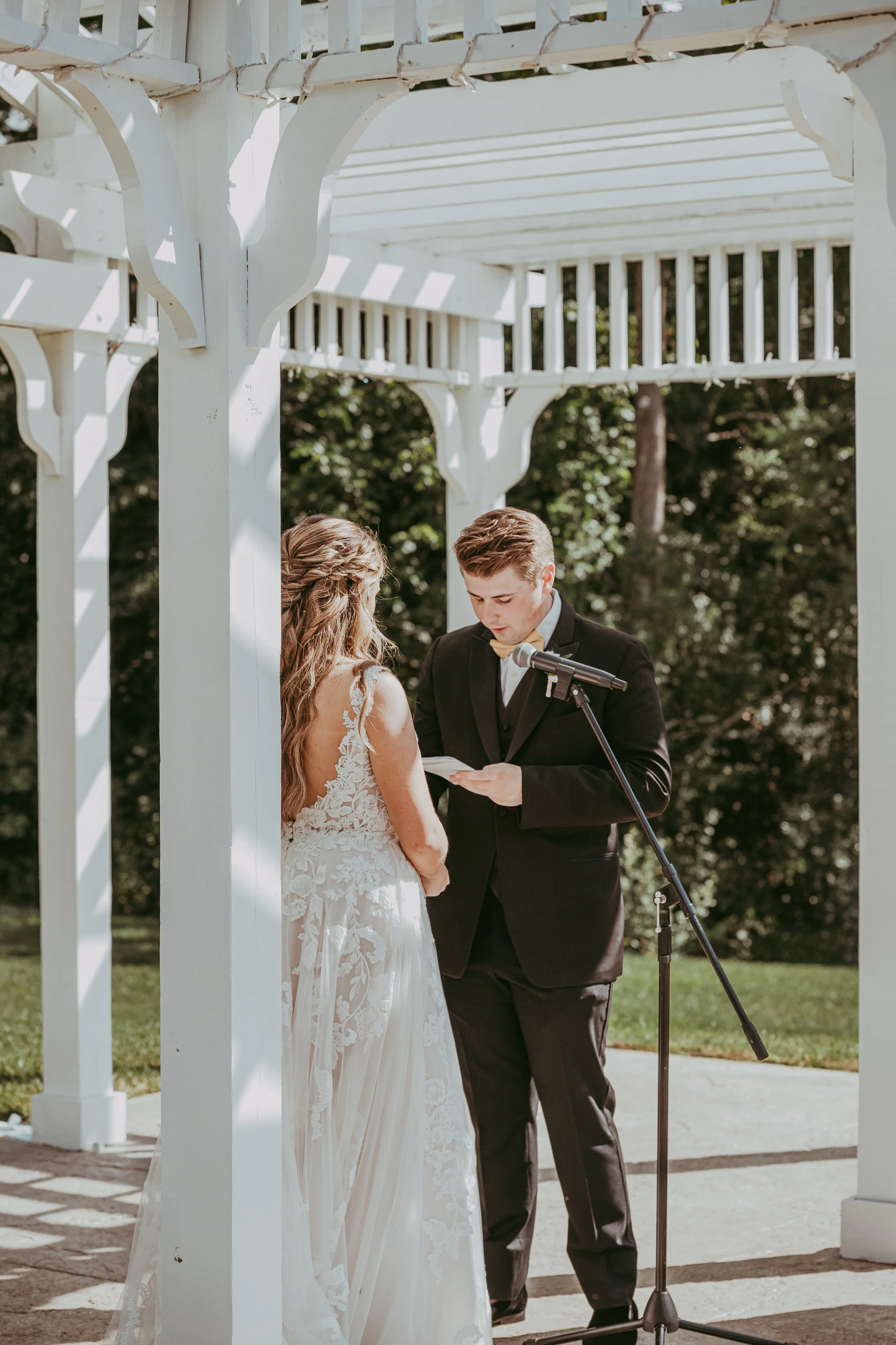 A couple getting married outdoors under a white wooden gazebo, with the groom reading vows from a paper, surrounded by greenery and bright sunlight.