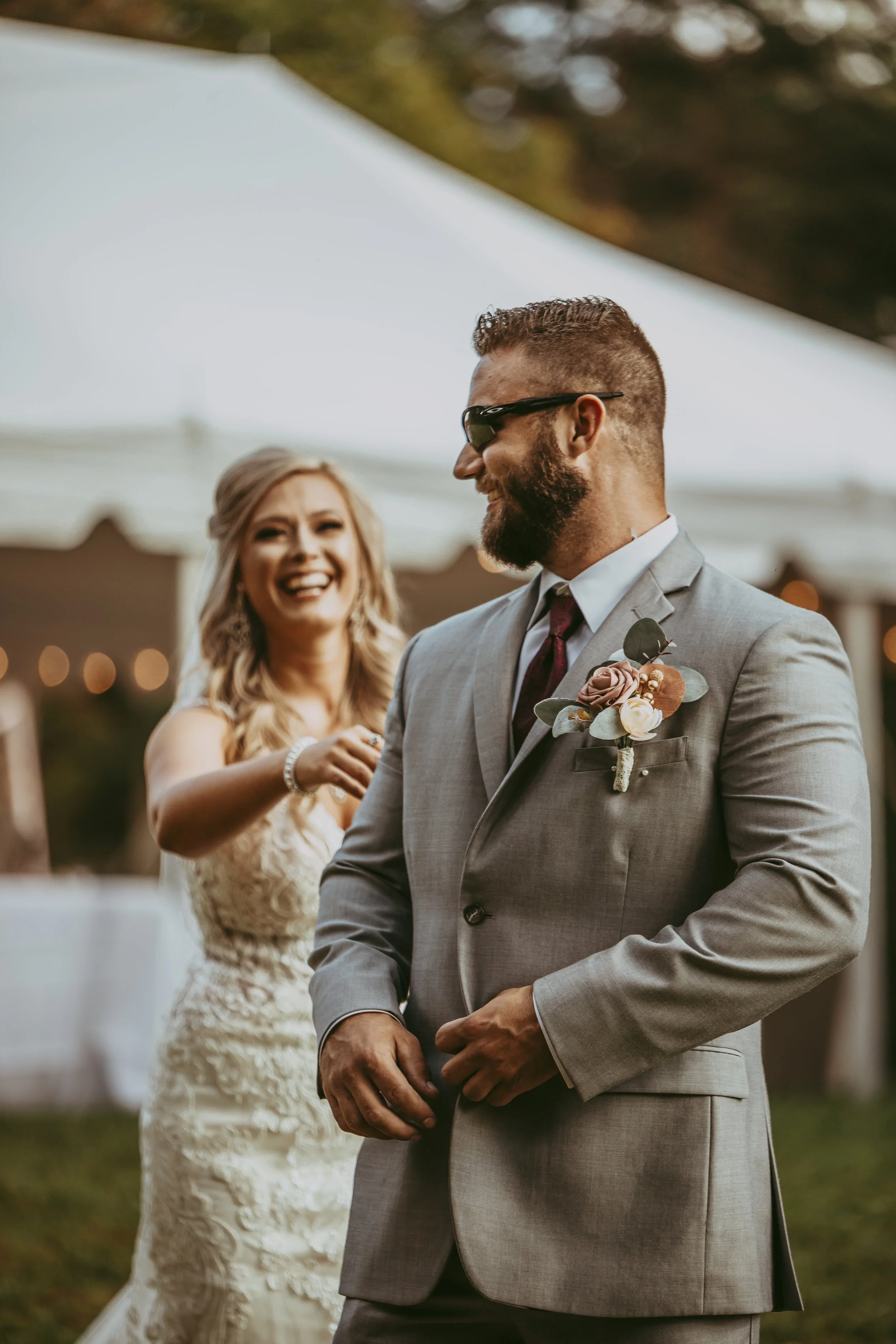 A bride and groom at a wedding reception outdoors, smiling and enjoying the moment. The groom wears a gray suit with a boutonniere, sunglasses, and a tie. The bride is in a lace wedding dress with curly blonde hair.