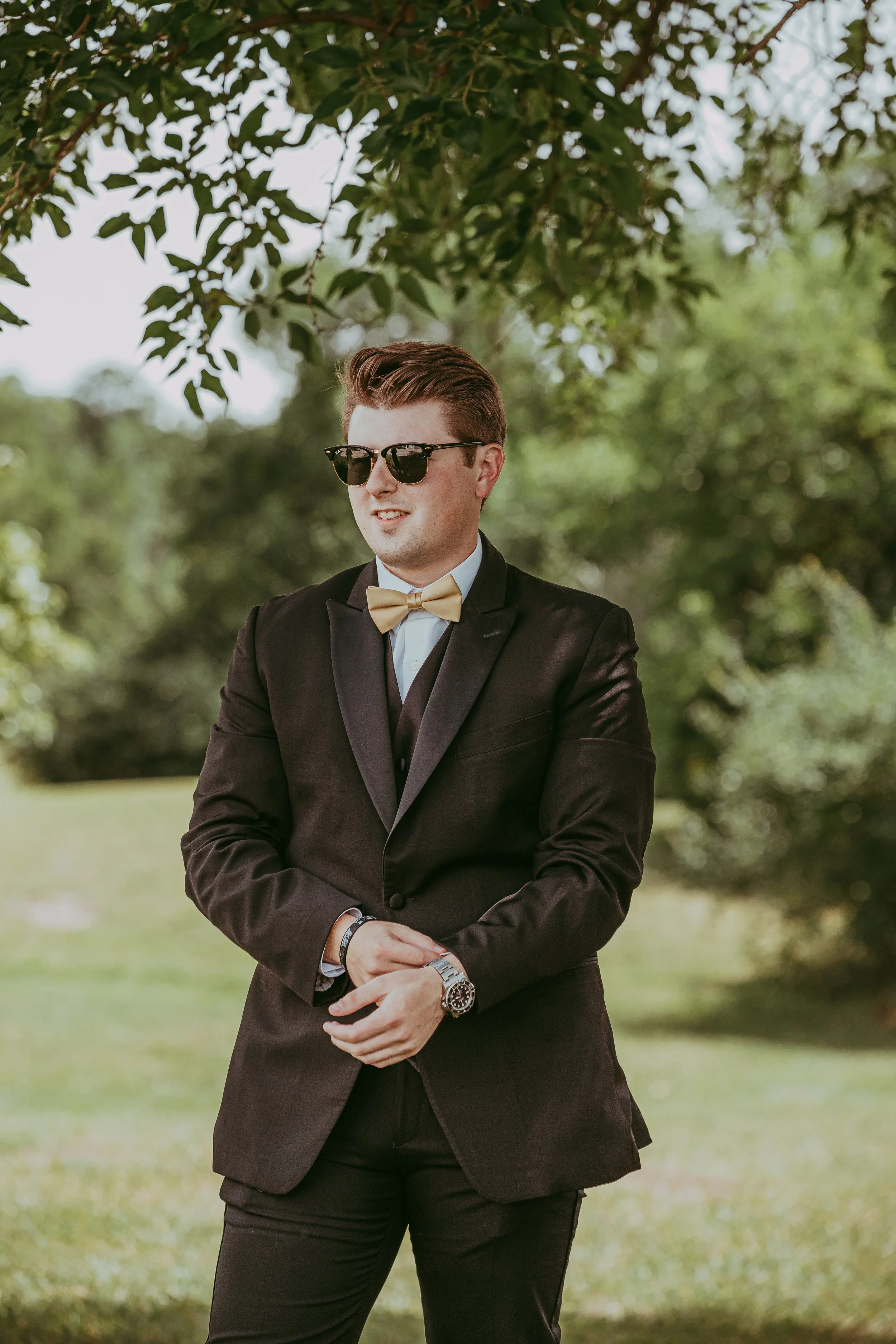 A man in a black suit with a beige bow tie and sunglasses standing outdoors under a tree.