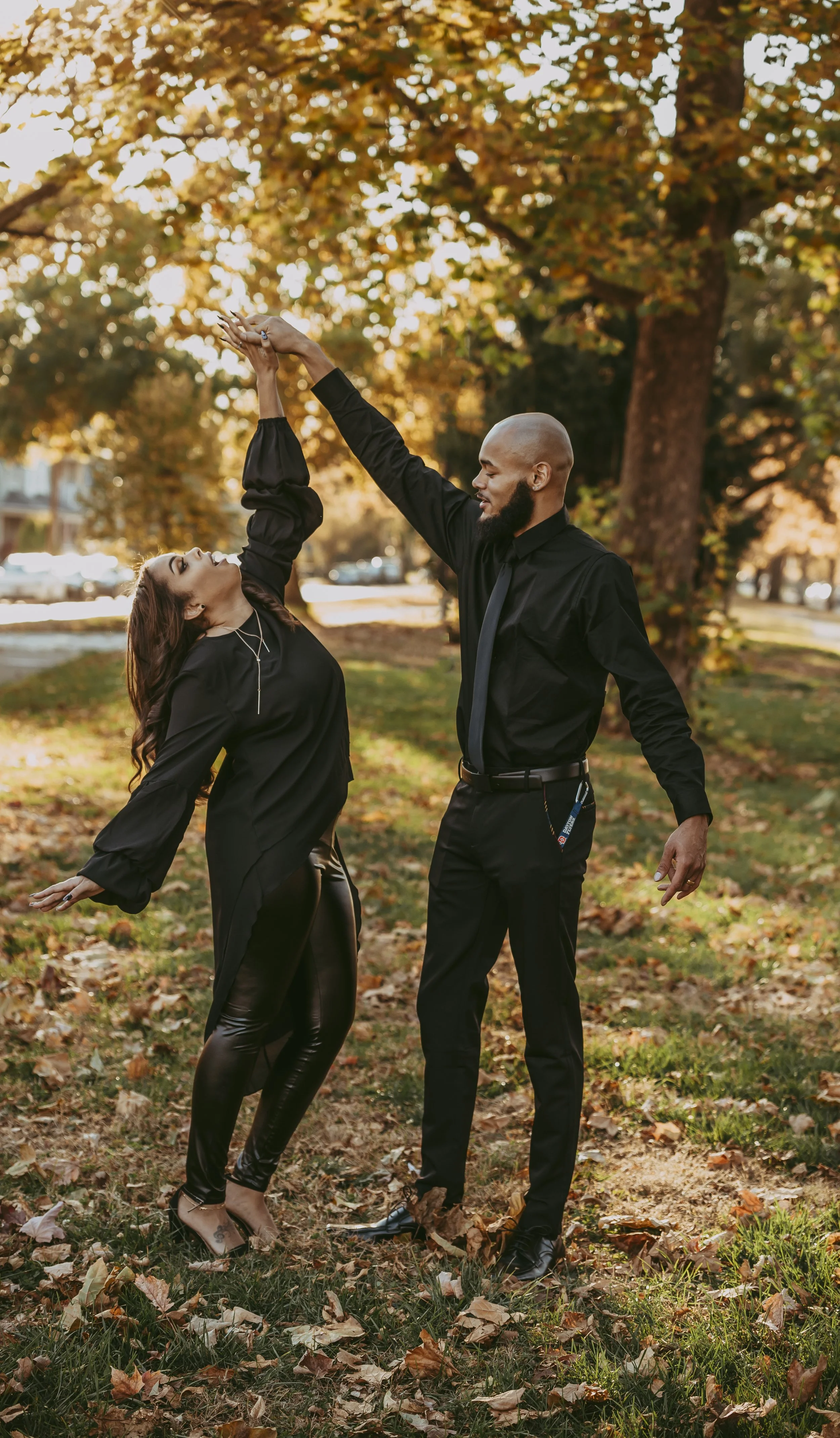 A man and woman dancing outdoors in a park during fall, with trees with autumn leaves in the background.