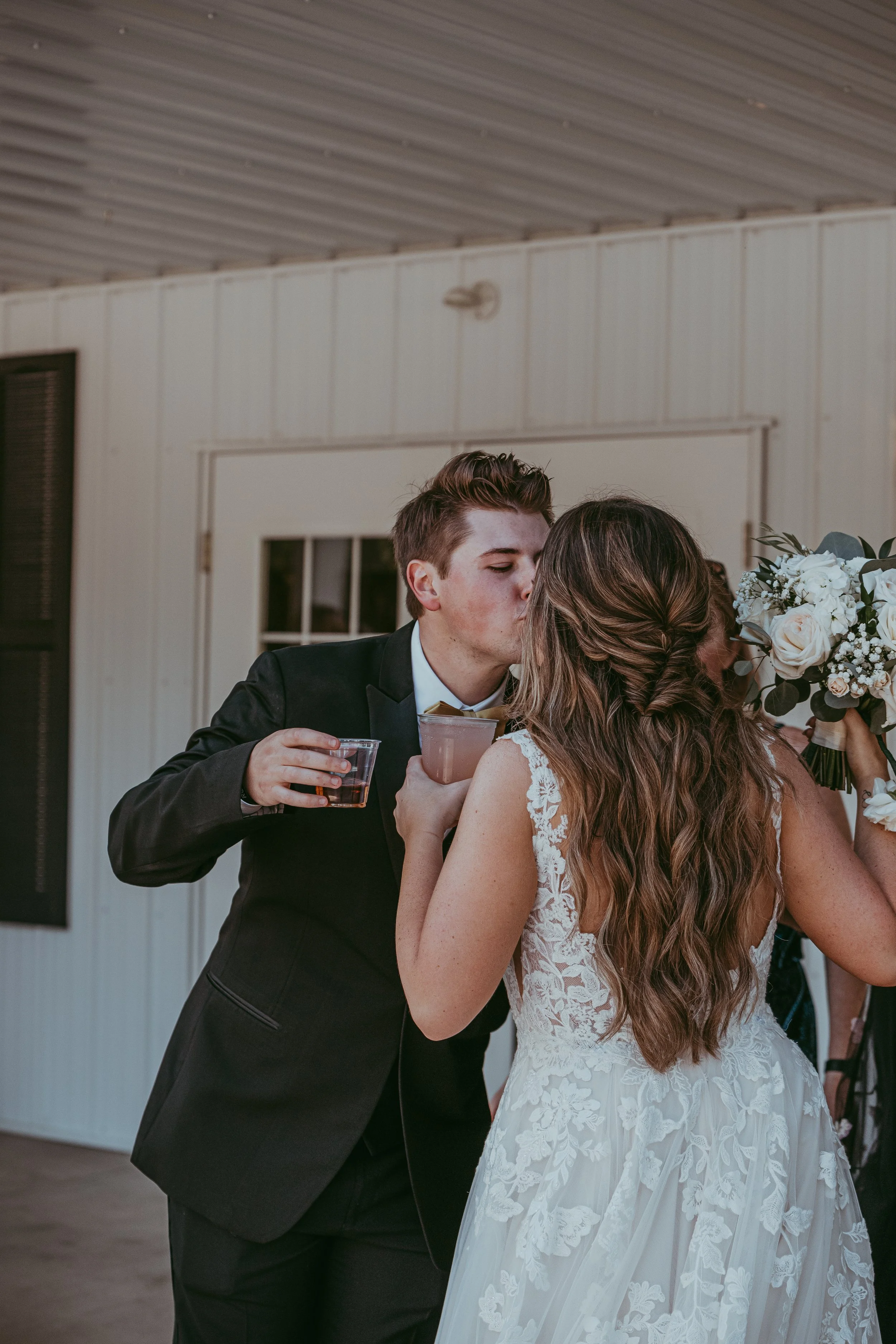 A young man in a tuxedo and a young woman in a wedding dress sharing a kiss during a wedding celebration, with the man holding drinks and the woman holding a bouquet of flowers.