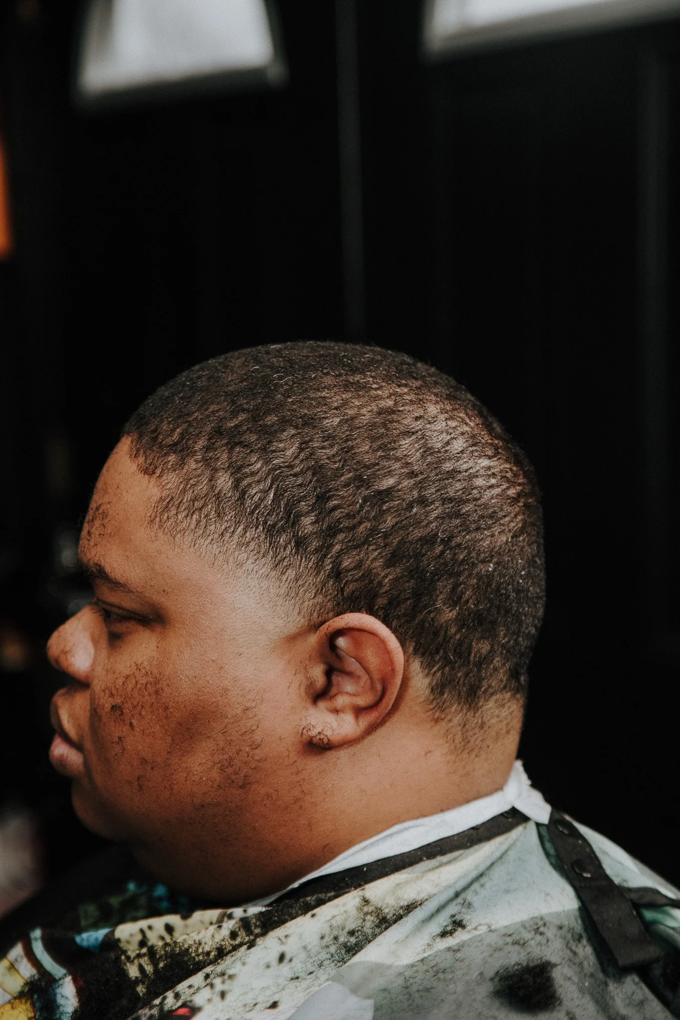 Side profile of a young man with short, textured black hair, sitting in a haircutting chair with a barber's cape, in a barbershop setting.