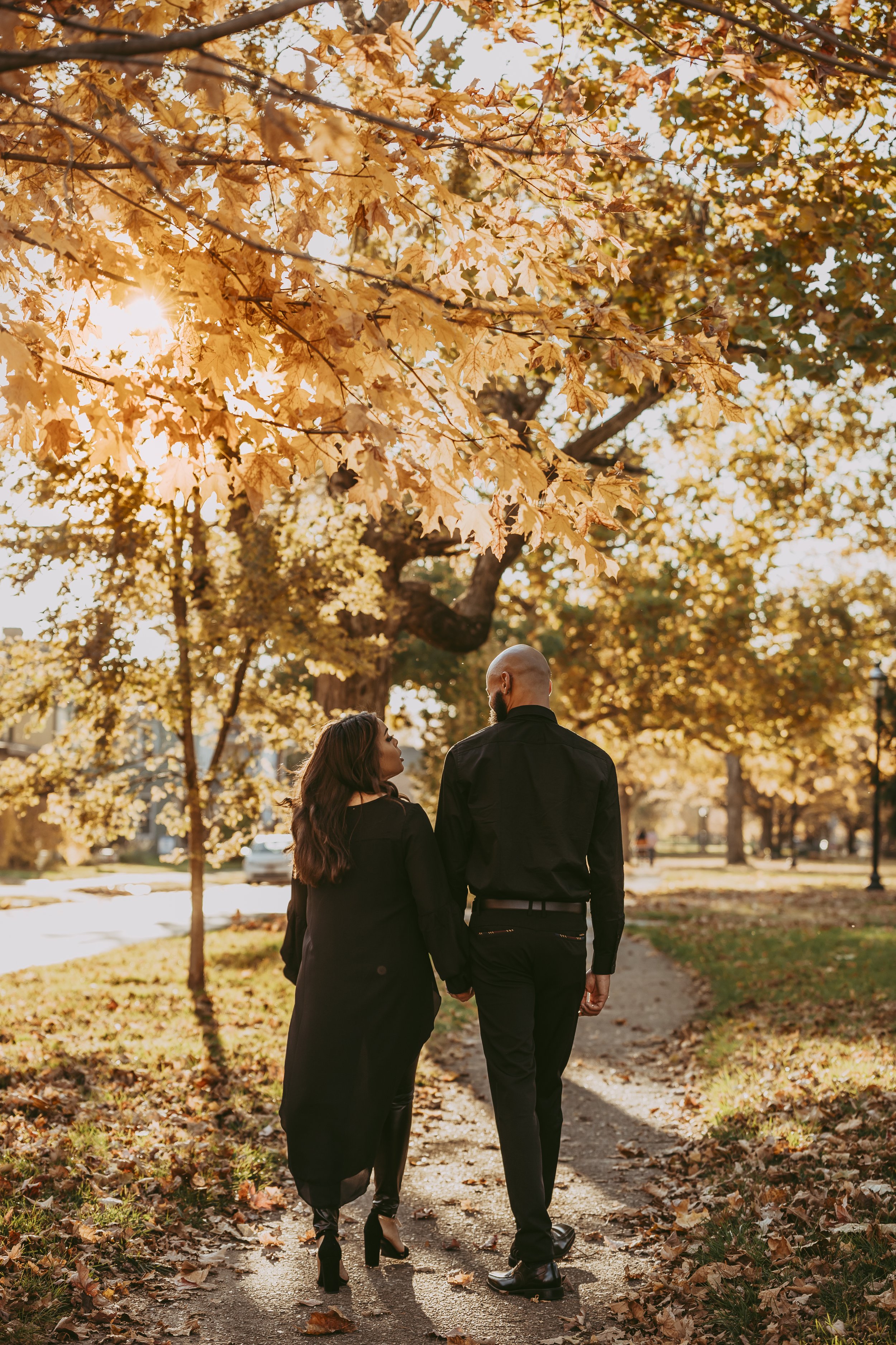 A couple walking hand in hand on a leaf-covered sidewalk during autumn, with golden sunlight filtering through colorful fall leaves overhead.