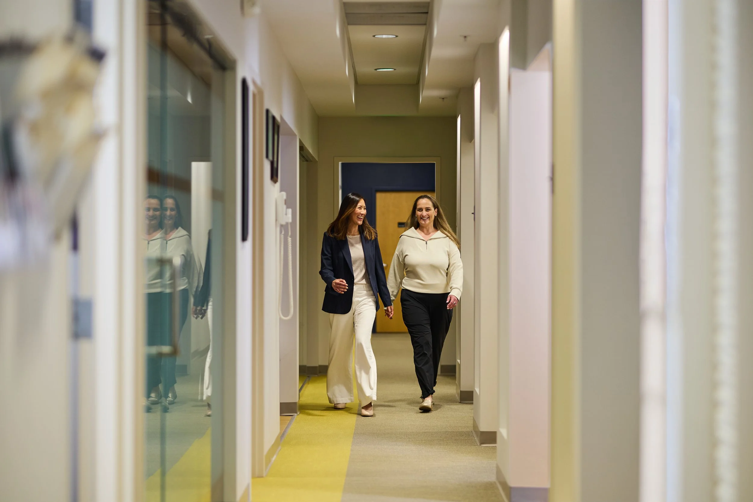 Two women walking down an office hallway, smiling and talking, dressed in business casual attire.