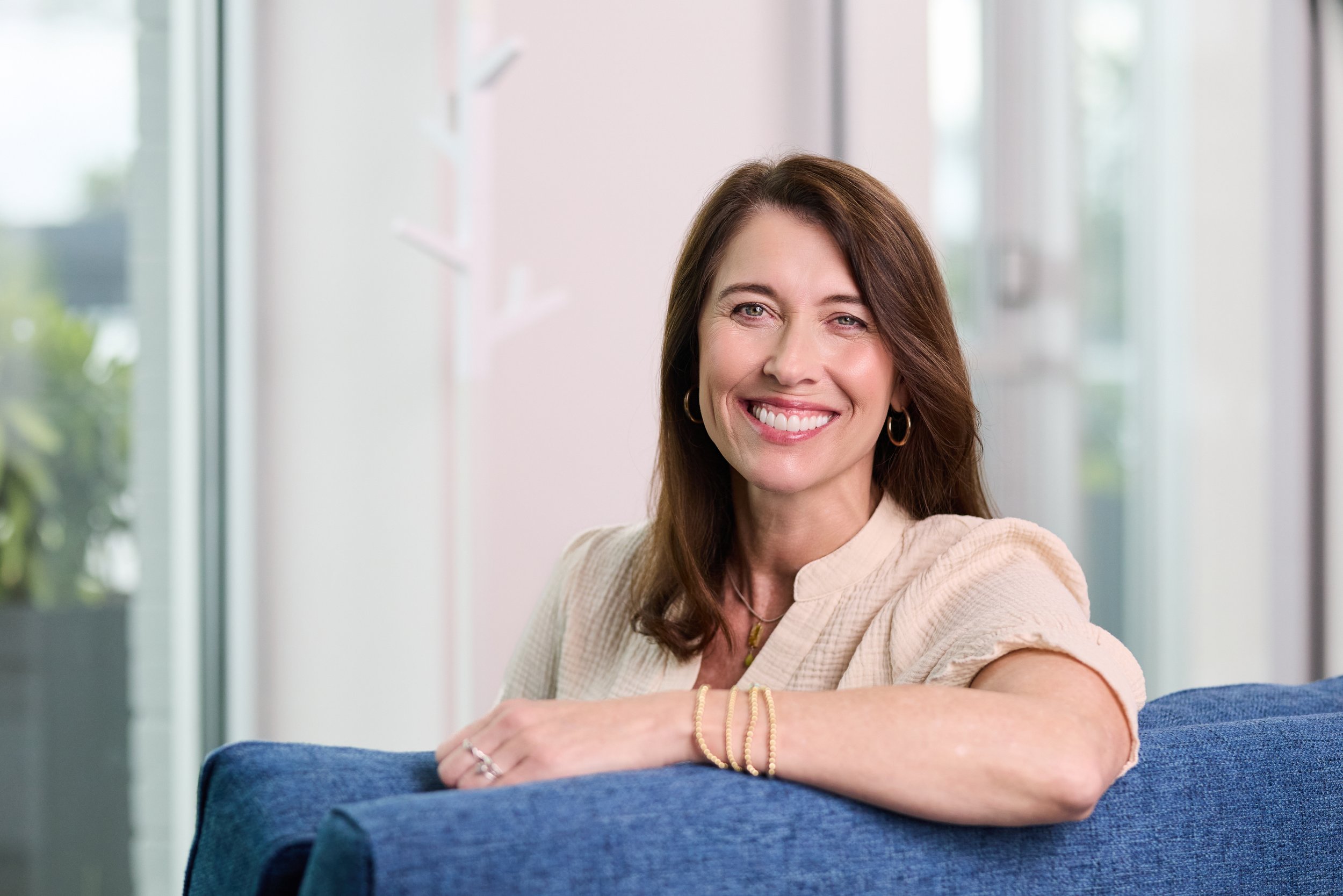 Smiling woman with brown hair and hoop earrings sitting on a blue couch in a bright room with large windows.