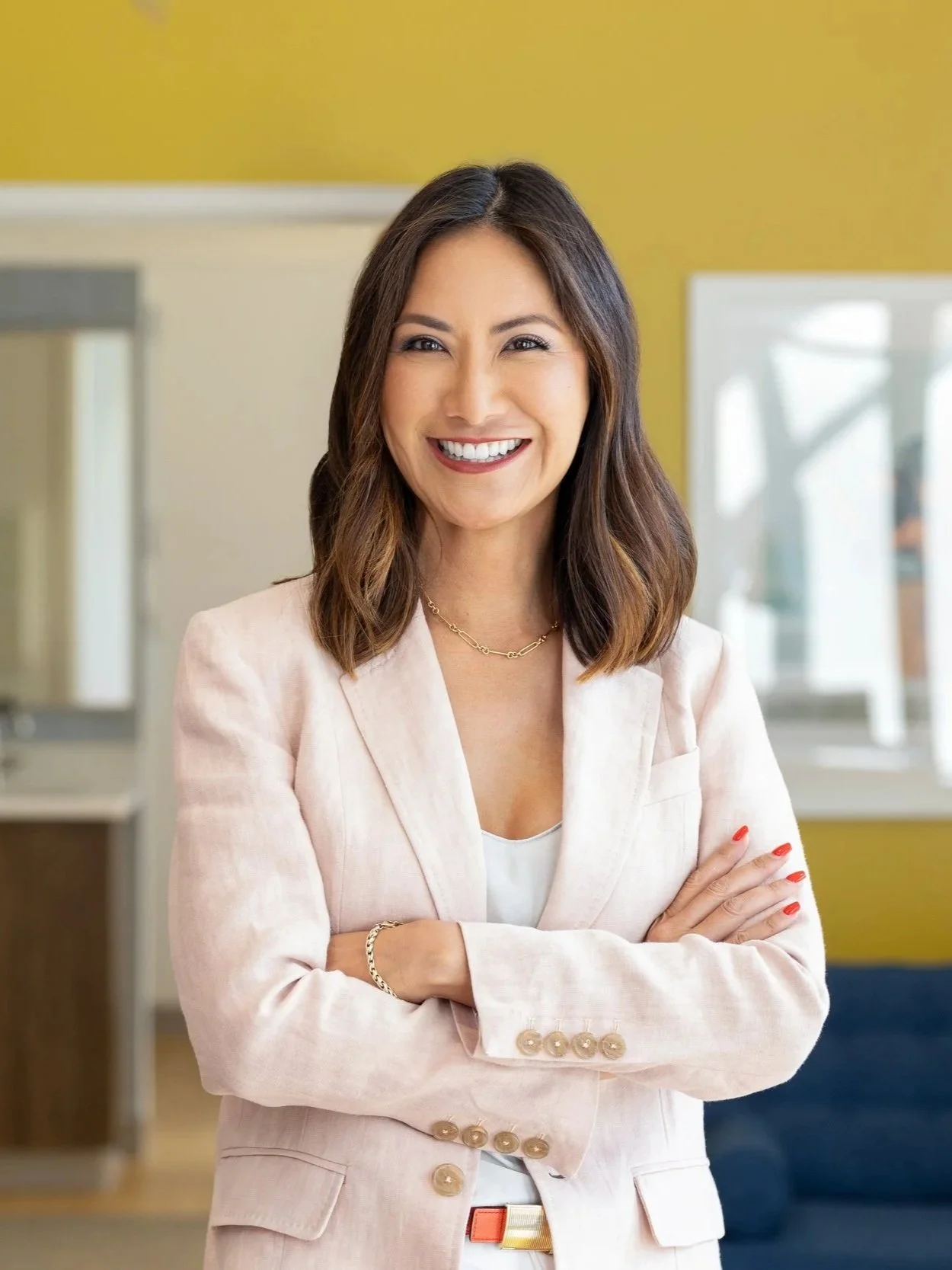 Woman with brown wavy hair standing and smiling in front of a yellow wall.