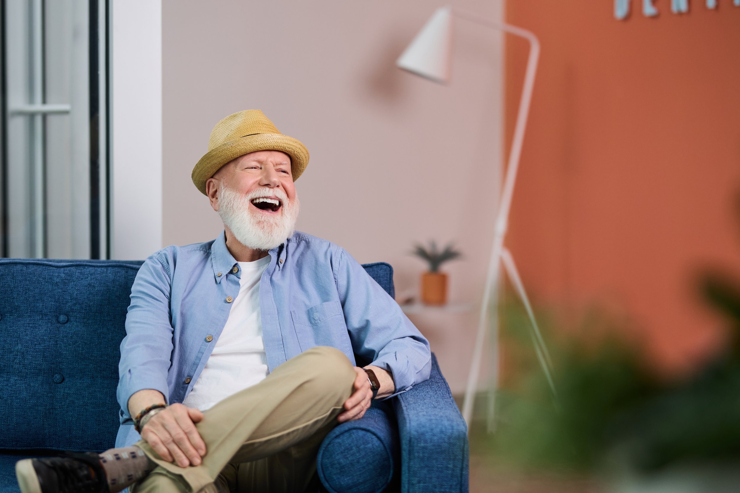 An elderly man with a beard and mustache wearing a tan hat, light blue button-up shirt, and khaki pants, sitting on a blue sofa, laughing while watching television in a brightly lit room.