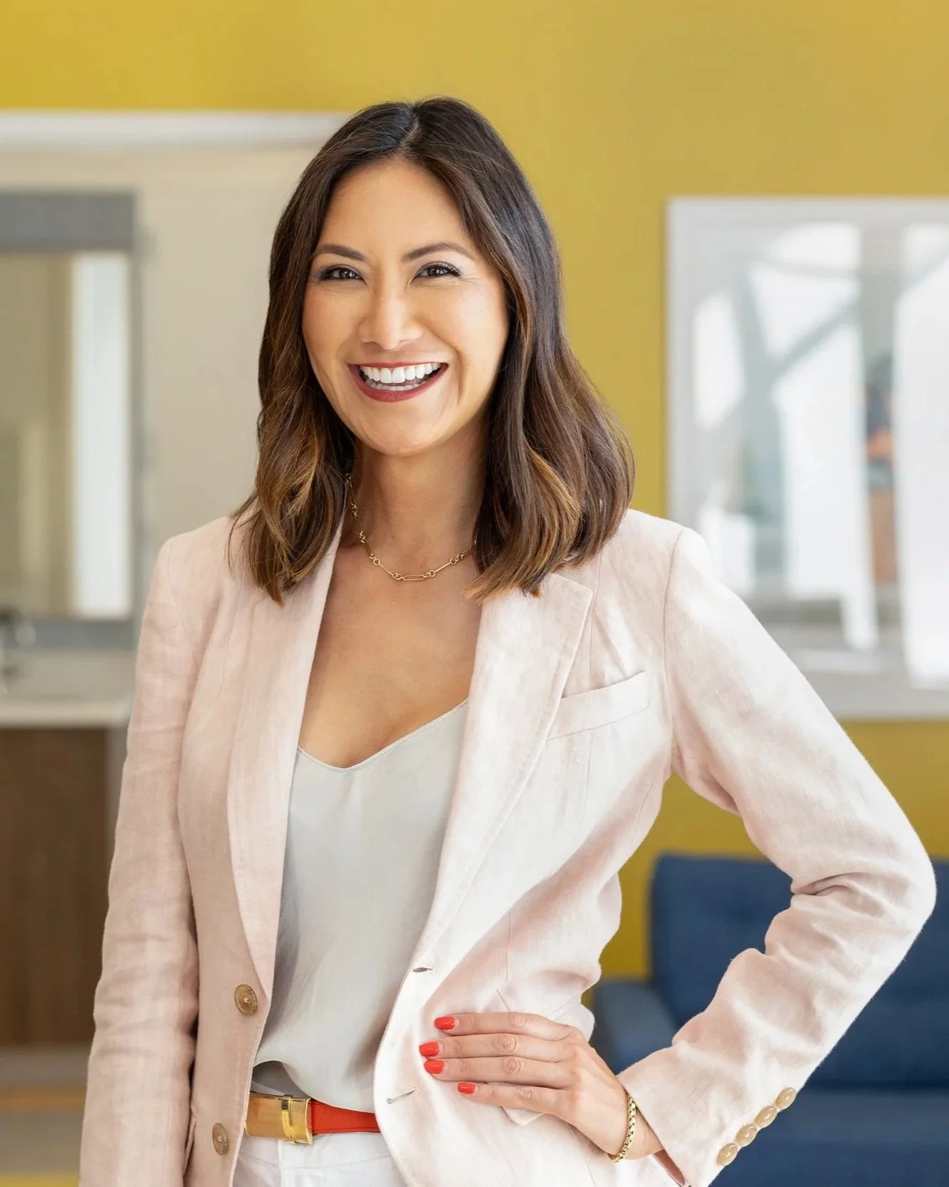 A woman with shoulder-length brown hair smiling, standing indoors with a yellow wall and reflective windows in the background, wearing a light pink blazer, white top, red belt, and jewelry.