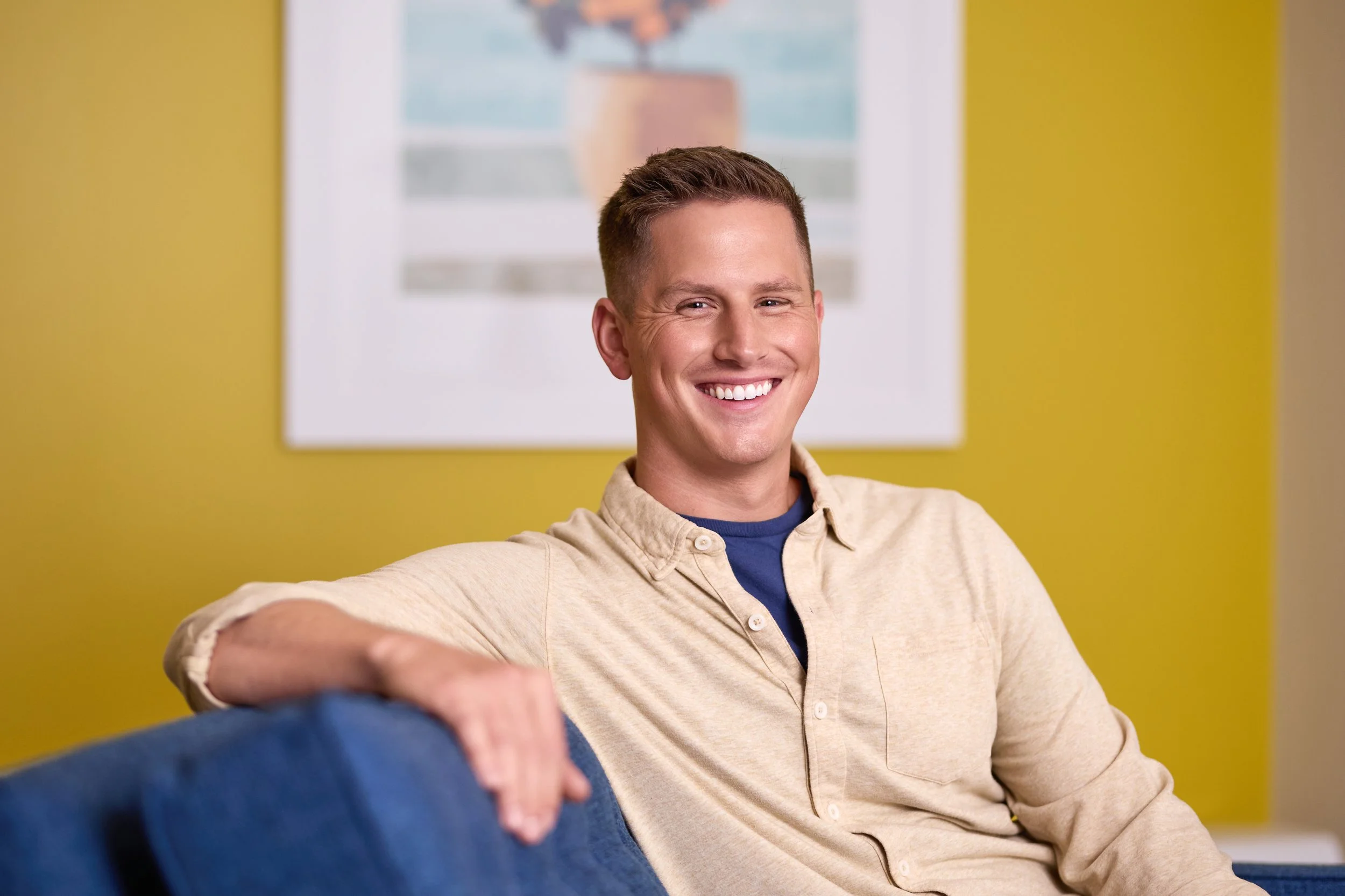 Smiling man sitting on a blue couch with a yellow wall and framed picture in the background.