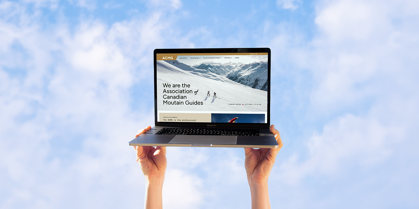 A person holding a laptop with a mountains guide website on the screen against a cloudy sky.