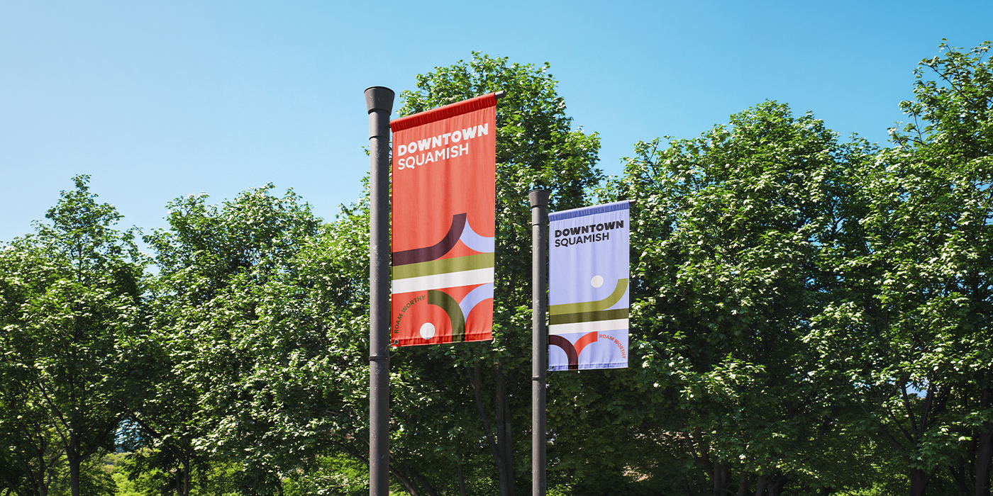 Two colorful vertical banners hanging from black poles with trees and a clear blue sky in the background. The banners read "Downtown Squamish."