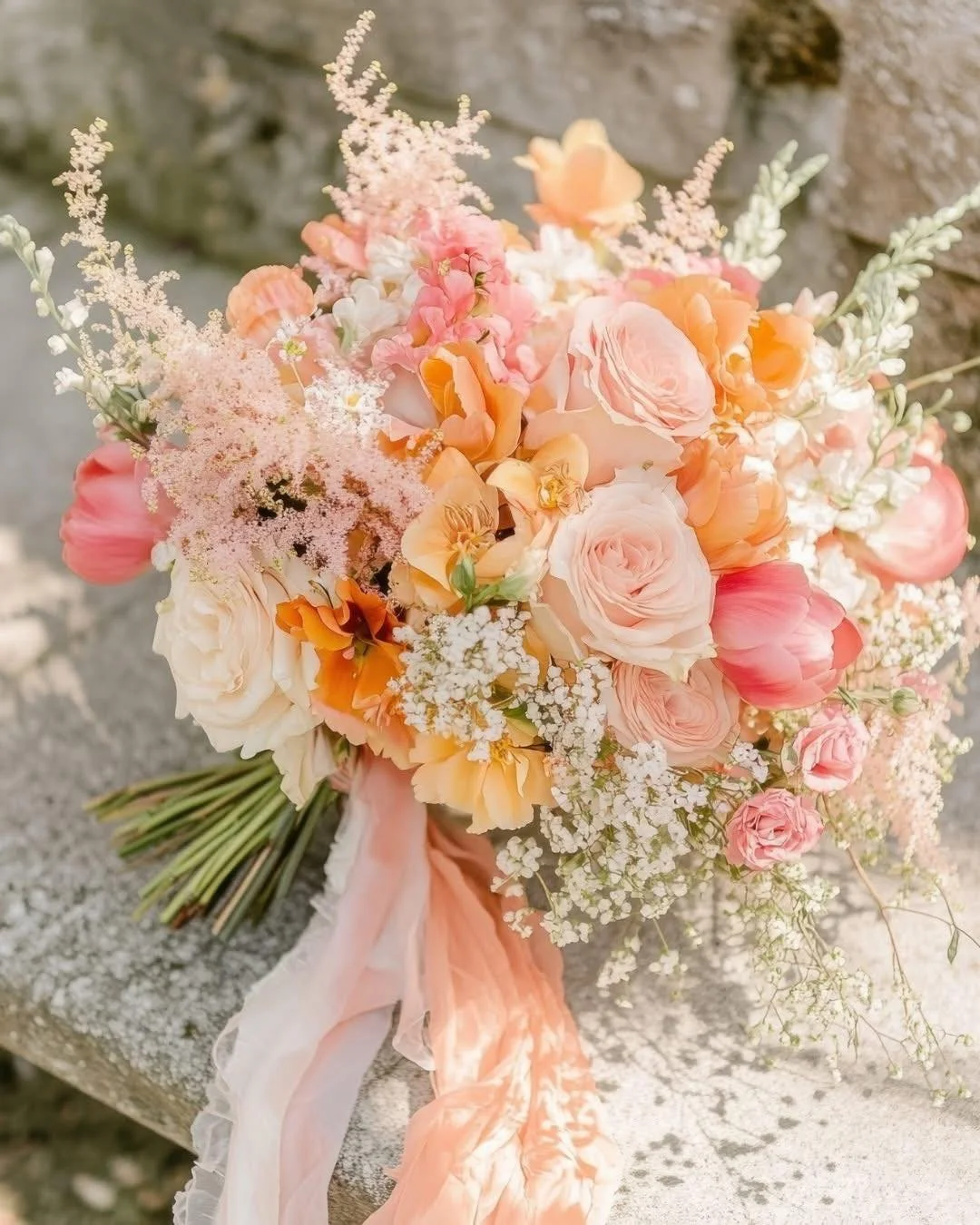 💕 Peachy roses and blush blooms mingle with wispy astilbe in this sun-kissed orange wedding bouquet. The vibrant accents add warmth while baby's breath creates airy volume. Those flowing silk ribbons add just the right touch of romance.