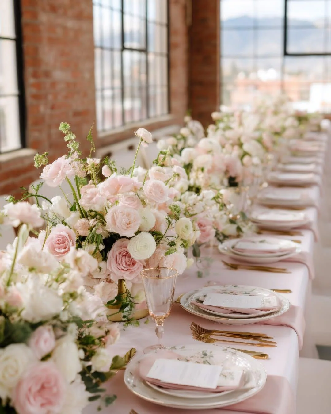 Soft pink roses and white ranunculus bring feminine charm to this industrial loft setting. The golden accents on the place settings complement the warm tones beautifully, proving that pastels work everywhere.