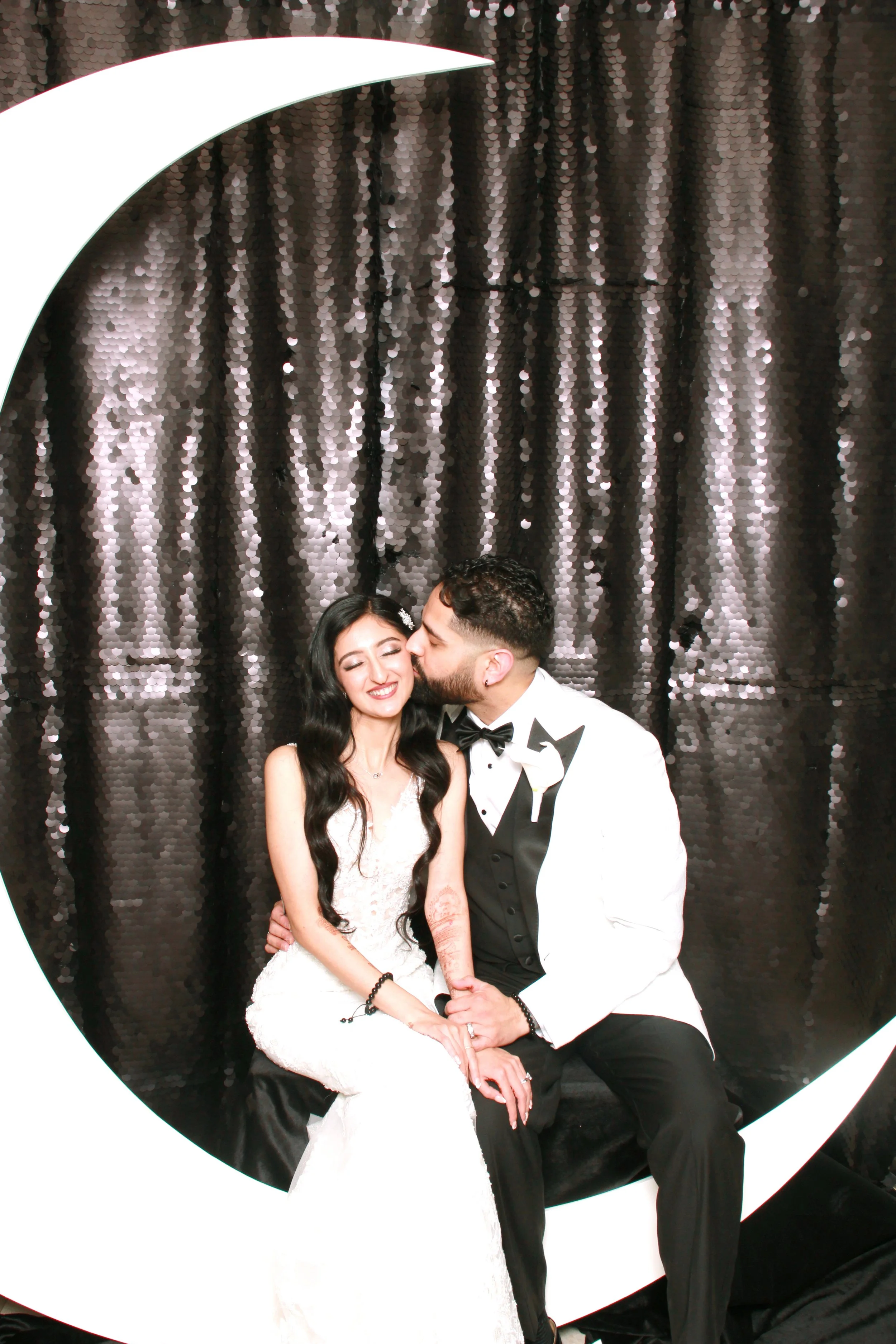 A happy bride and groom sitting inside a large white crescent moon shape, with the groom kissing the bride on the cheek, in front of a dark, sequined backdrop.
