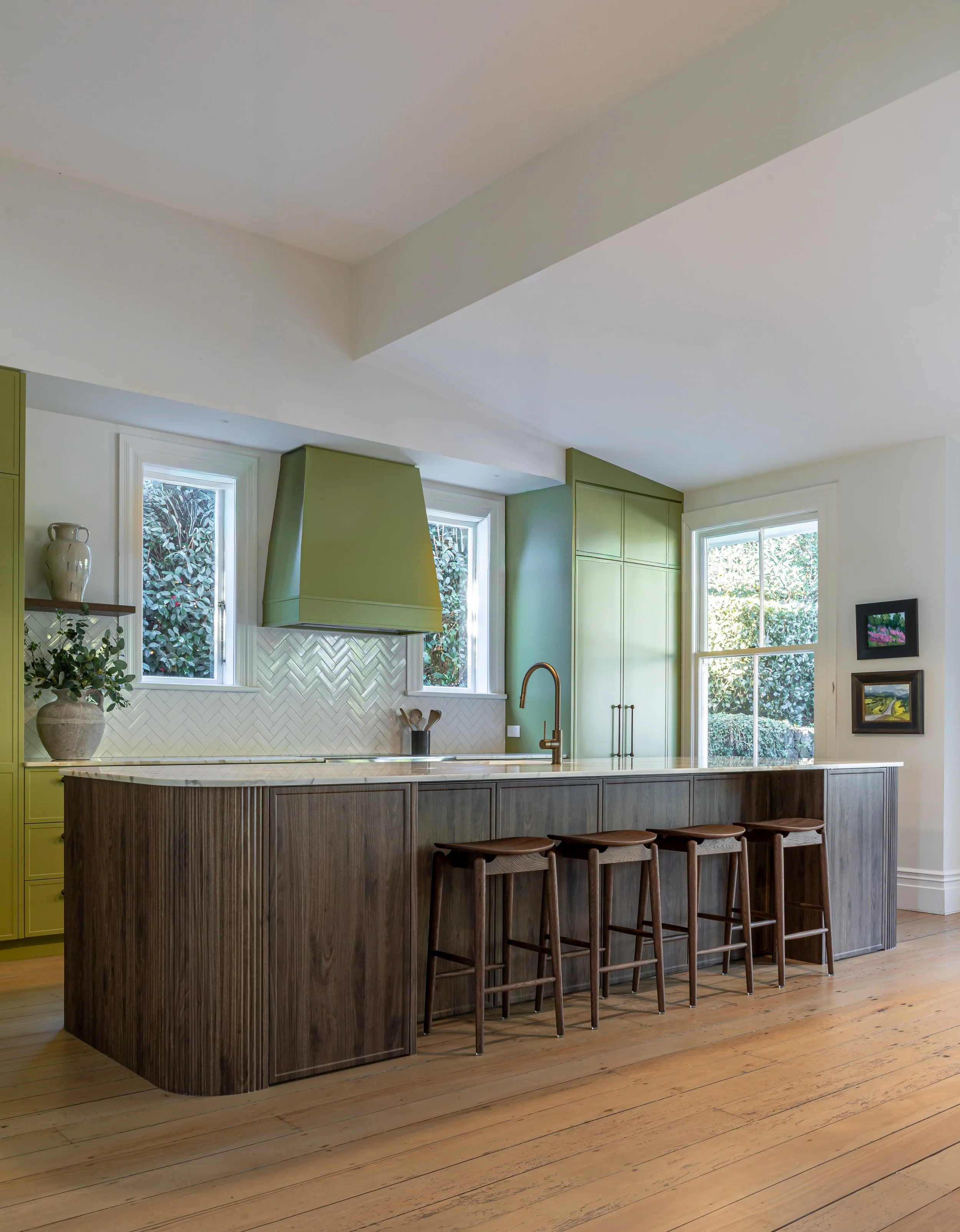 Modern kitchen with white cabinets, beige tiled backsplash, a marble island with bar stools, and large windows with greenery outside.