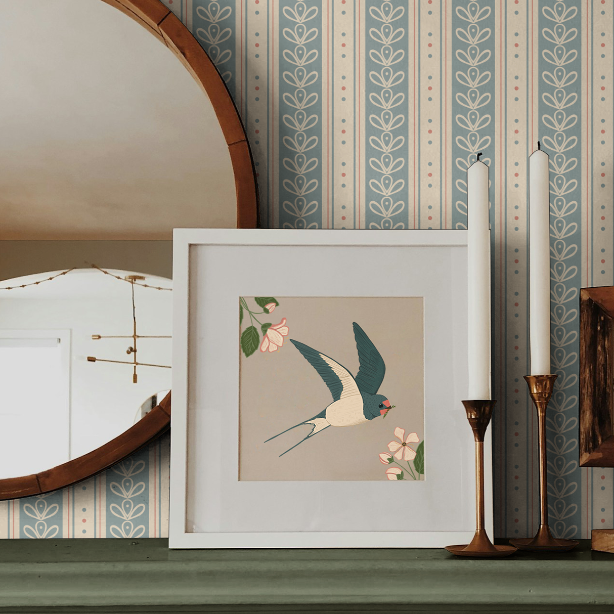 A decorative shelf with a framed illustration of a bird surrounded by pink flowers, two white candles in brass holders, and a mirrored wall with a wooden frame mirror in the background.