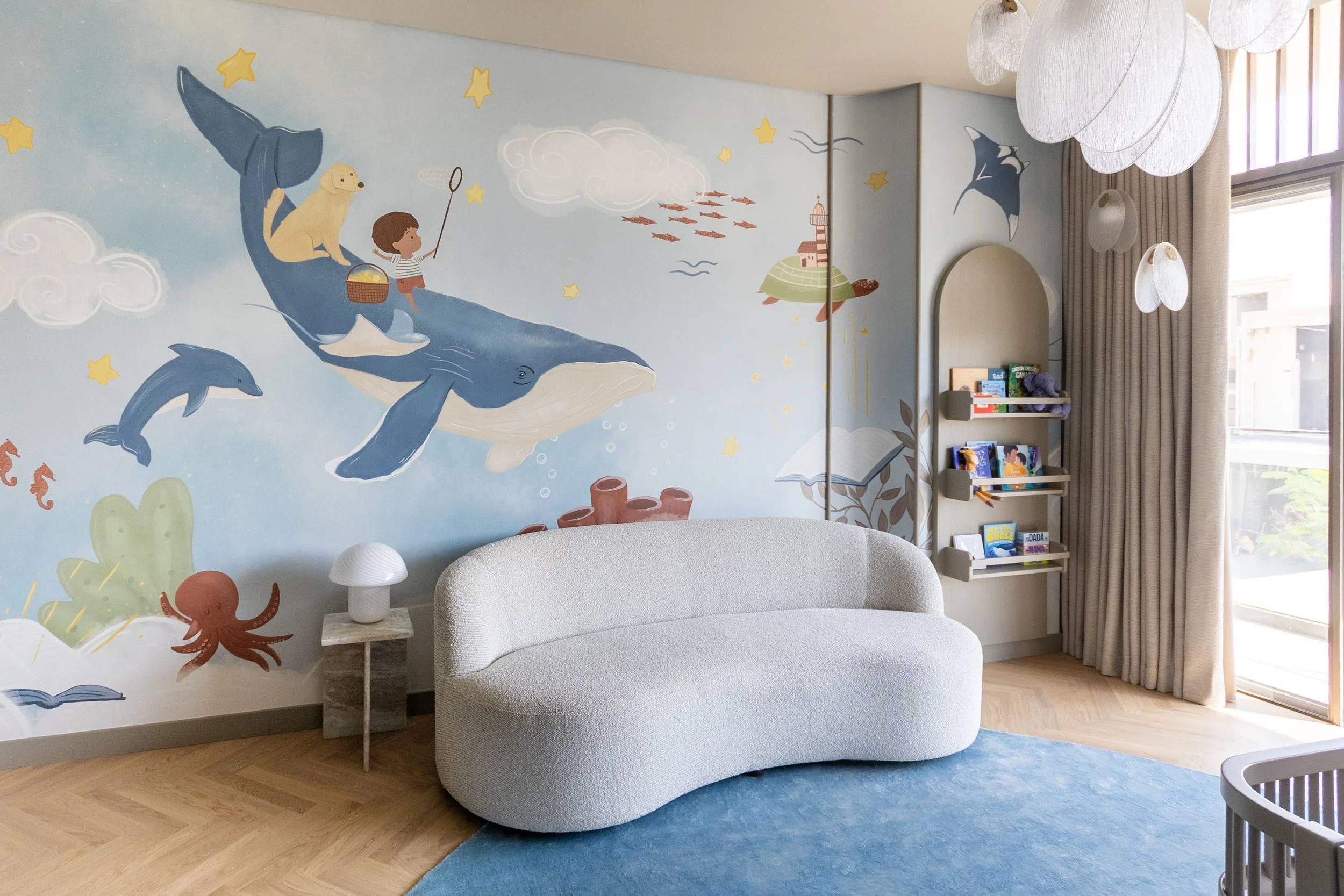 Nursery with ocean-themed wall mural featuring a boy riding a whale with a dog, dolphins, stars, clouds, an octopus, and fish, with a cozy beige sofa, bookshelf, and large window with curtains.