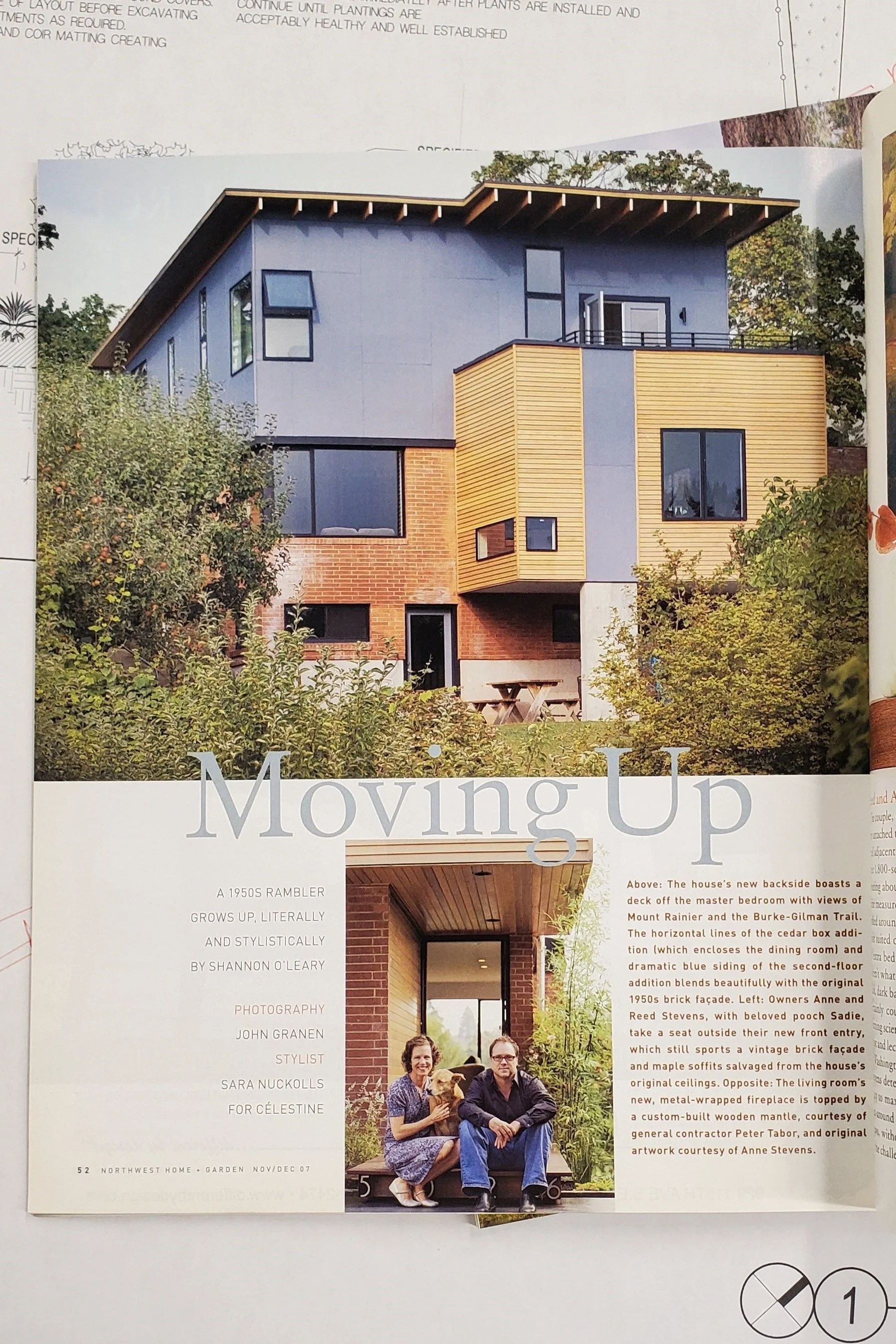 Magazine page featuring a modern house with wooden and blue siding, surrounded by greenery. The headline reads 'Moving Up.' Below the image is a section about the house with text describing its features. A smaller photo shows two people sitting with a dog, identified as house owners in the text.