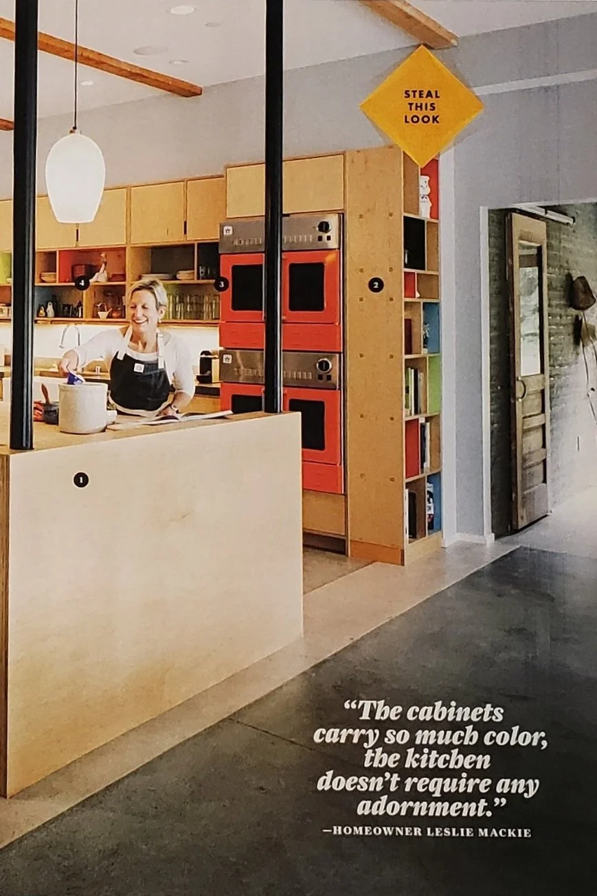 Modern kitchen with colorful cabinets and integrated red double ovens. A person in an apron is preparing food. Shelves with books and dishes are visible. Quote about kitchen decor by homeowner Leslie Mackie reads: 'The cabinets carry so much color, the kitchen doesn’t require any adornment.' A 'Steal This Look' sign is on the wall.