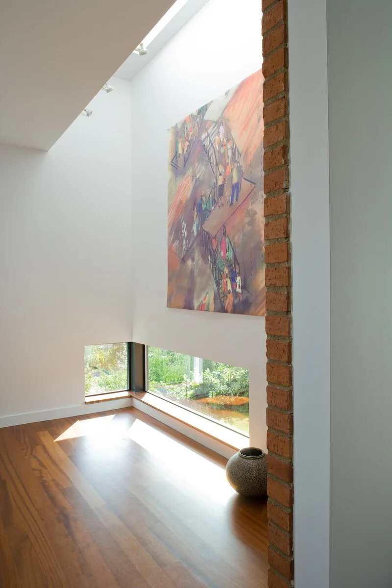 Interior of a modern room with a large painting on the wall, featuring abstract figures and stairs. The room has wooden flooring, a large window offering natural light, and a decorative vase.