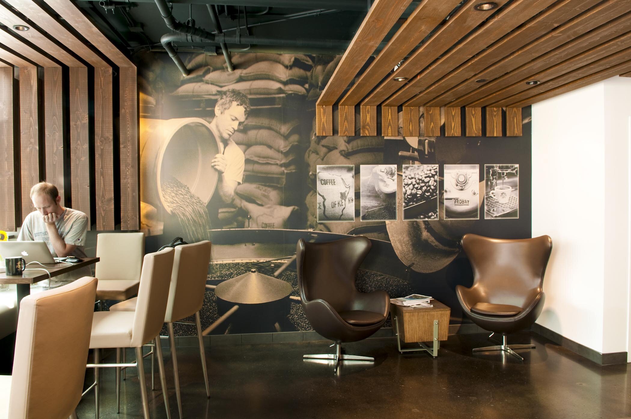 Coffee shop interior with modern chairs, wooden ceiling, and wall art depicting coffee processing.