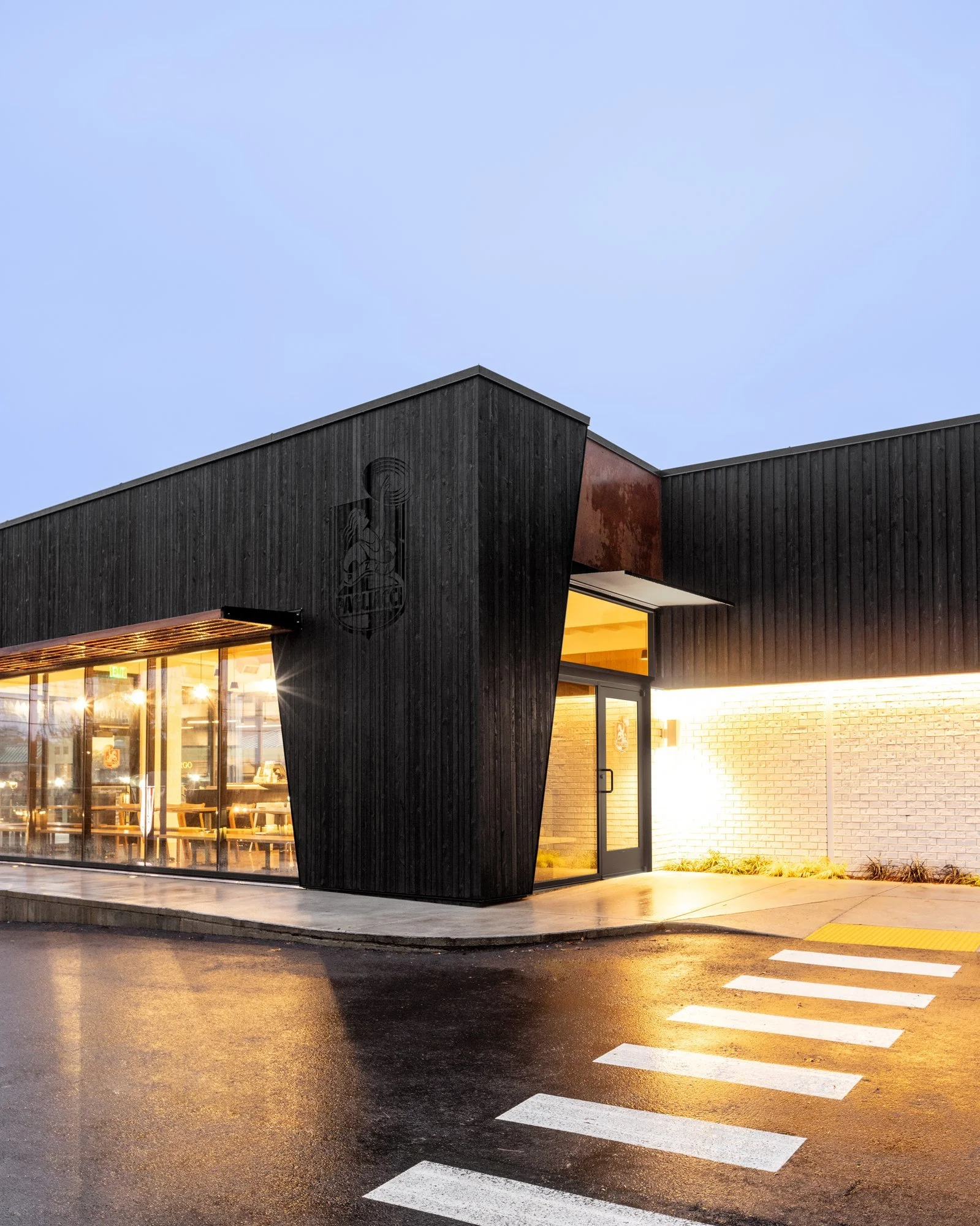 Modern black building exterior with large windows and illuminated interior, adjacent to a wet sidewalk and a crosswalk at dusk.