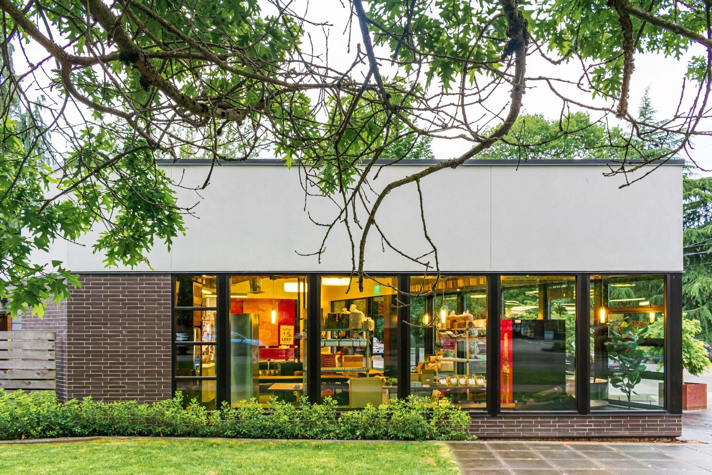 Modern building with large windows showcasing a brightly lit interior, surrounded by trees and a grassy area.