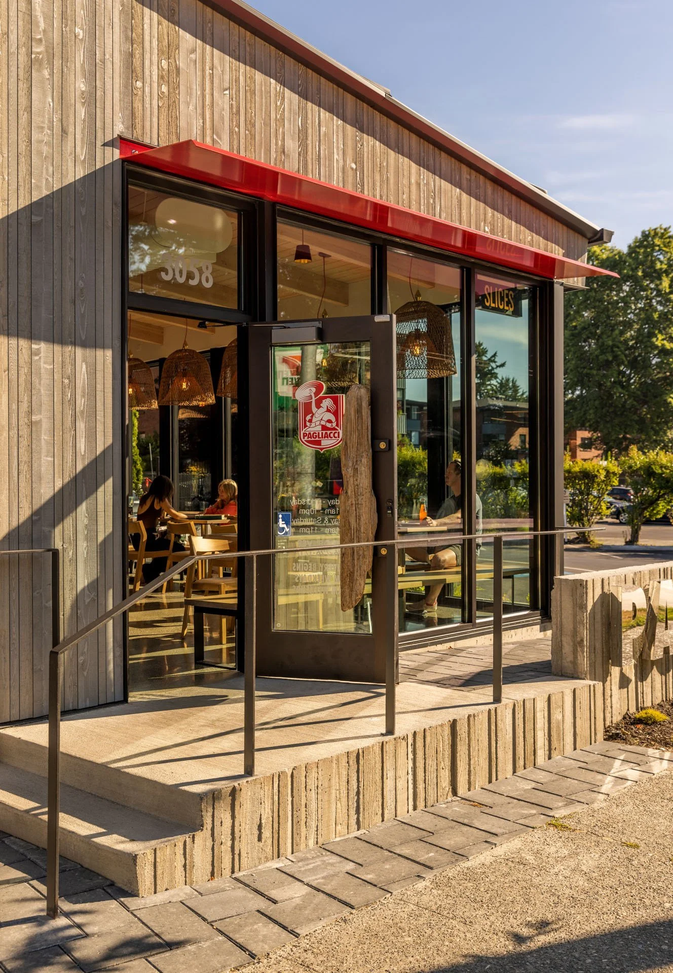 Exterior of a modern pizza restaurant with wood-paneling, glass door, and red awning, displaying 'Pagliacci Pizza' logo. Inside, people are visible dining.