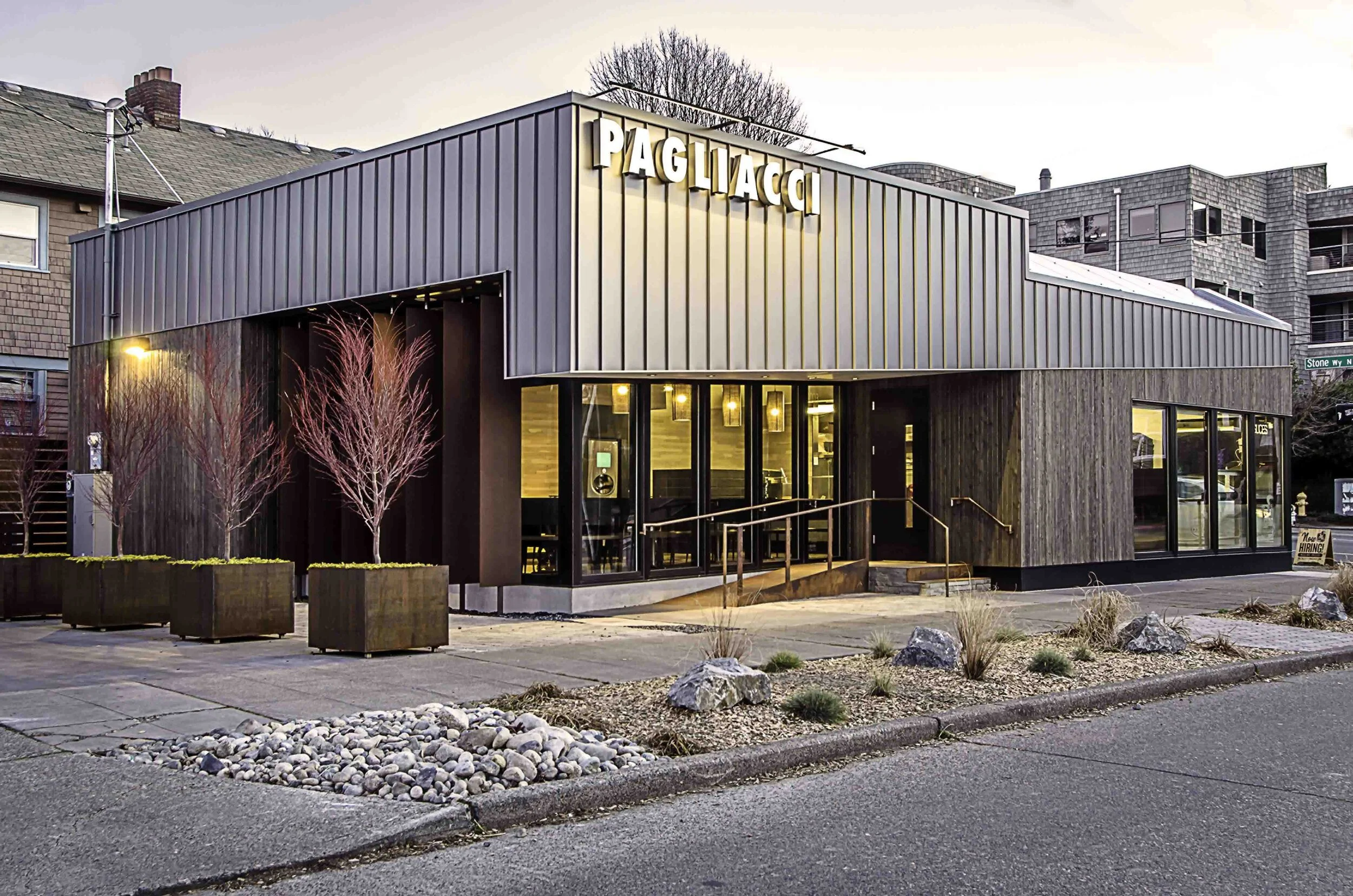 Modern building exterior with large windows and minimalist landscaping, displaying "PAGLIACCI" in illuminated letters at dusk.