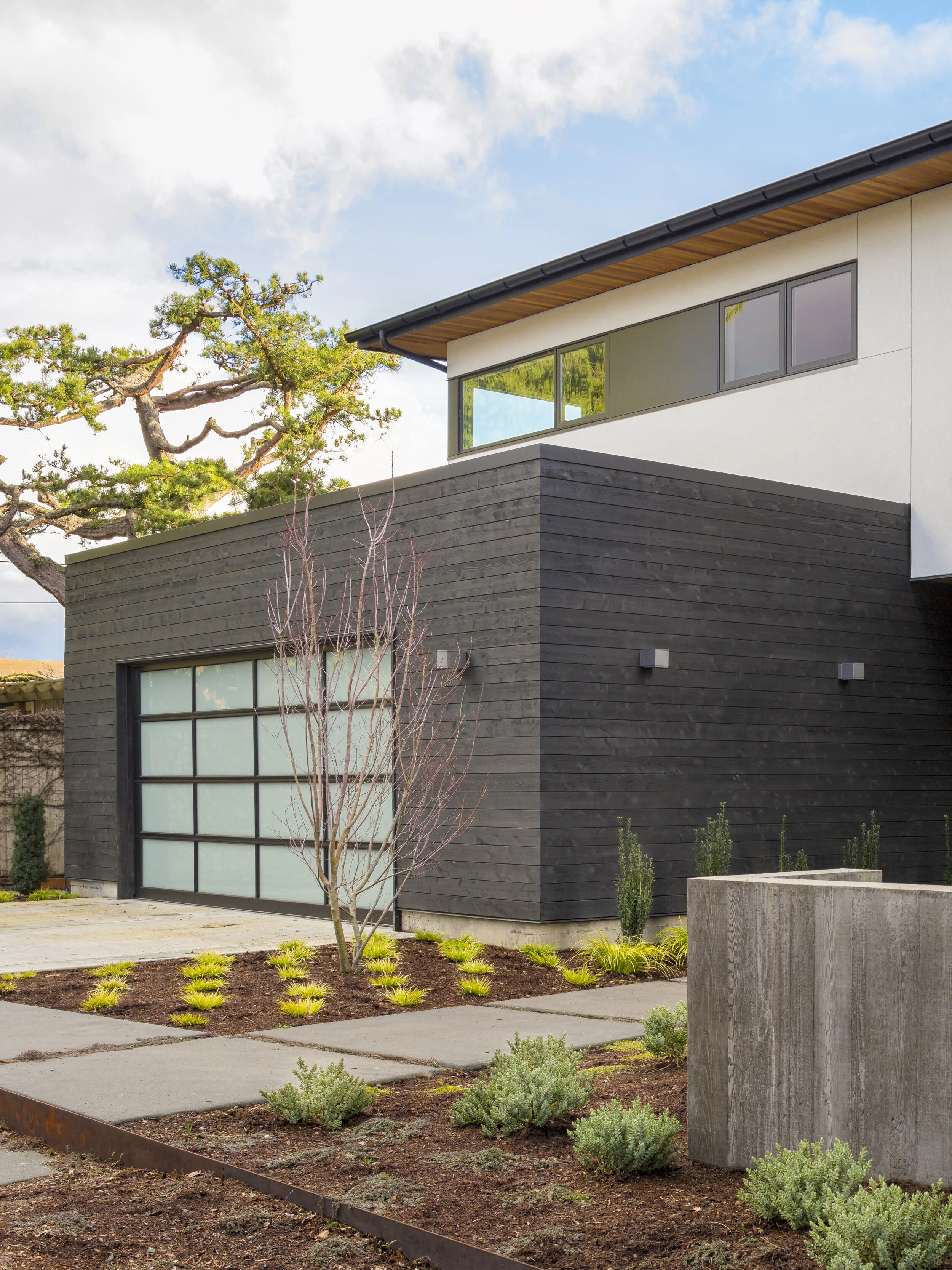 Modern house with dark wood siding and a glass garage door, featuring minimal landscaping and a tree against a cloudy blue sky.
