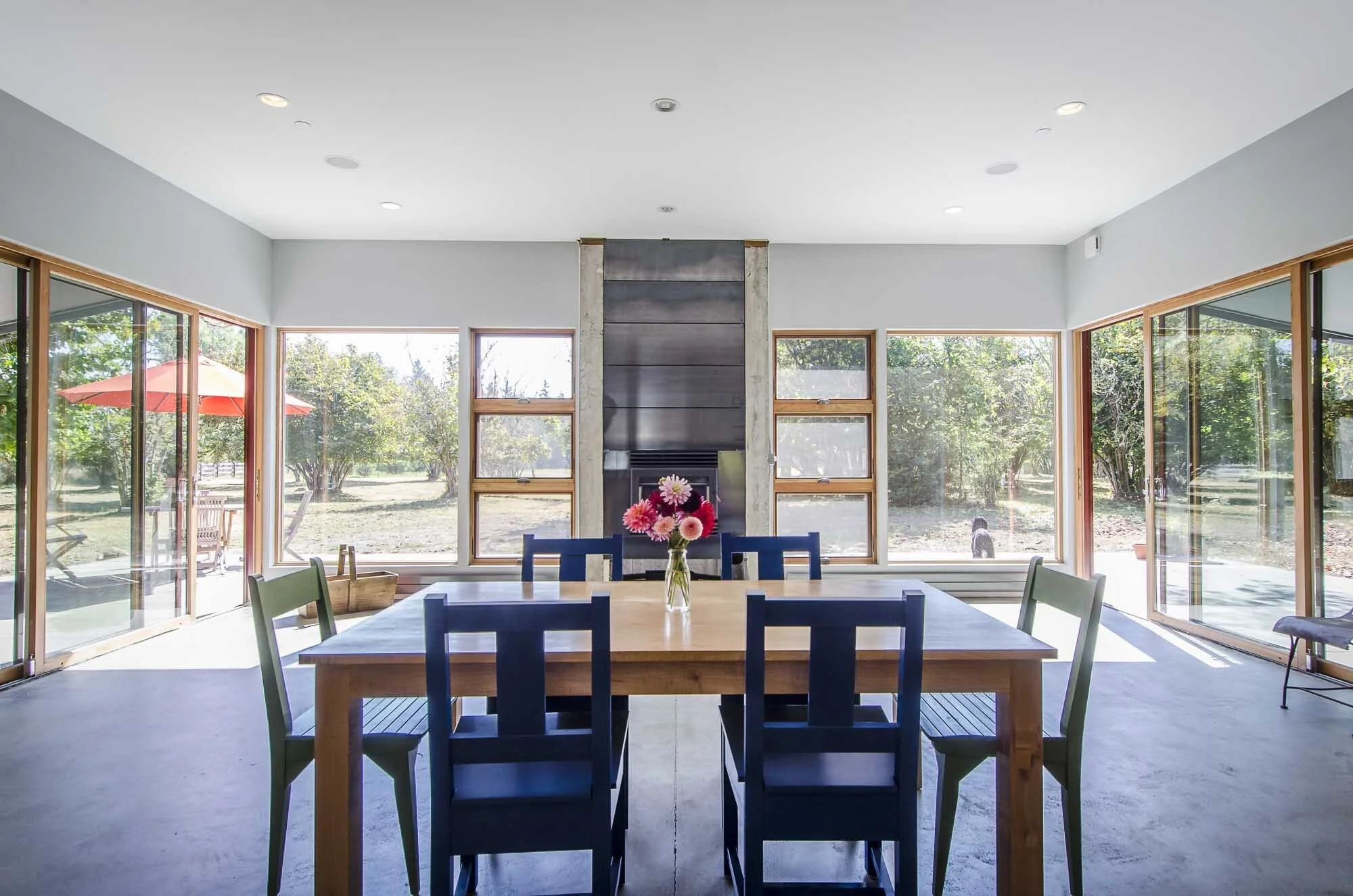 Modern dining room with a wooden table and blue chairs, featuring a vase of flowers in the center. Large windows offer views of a green outdoor area with trees and a red patio umbrella.