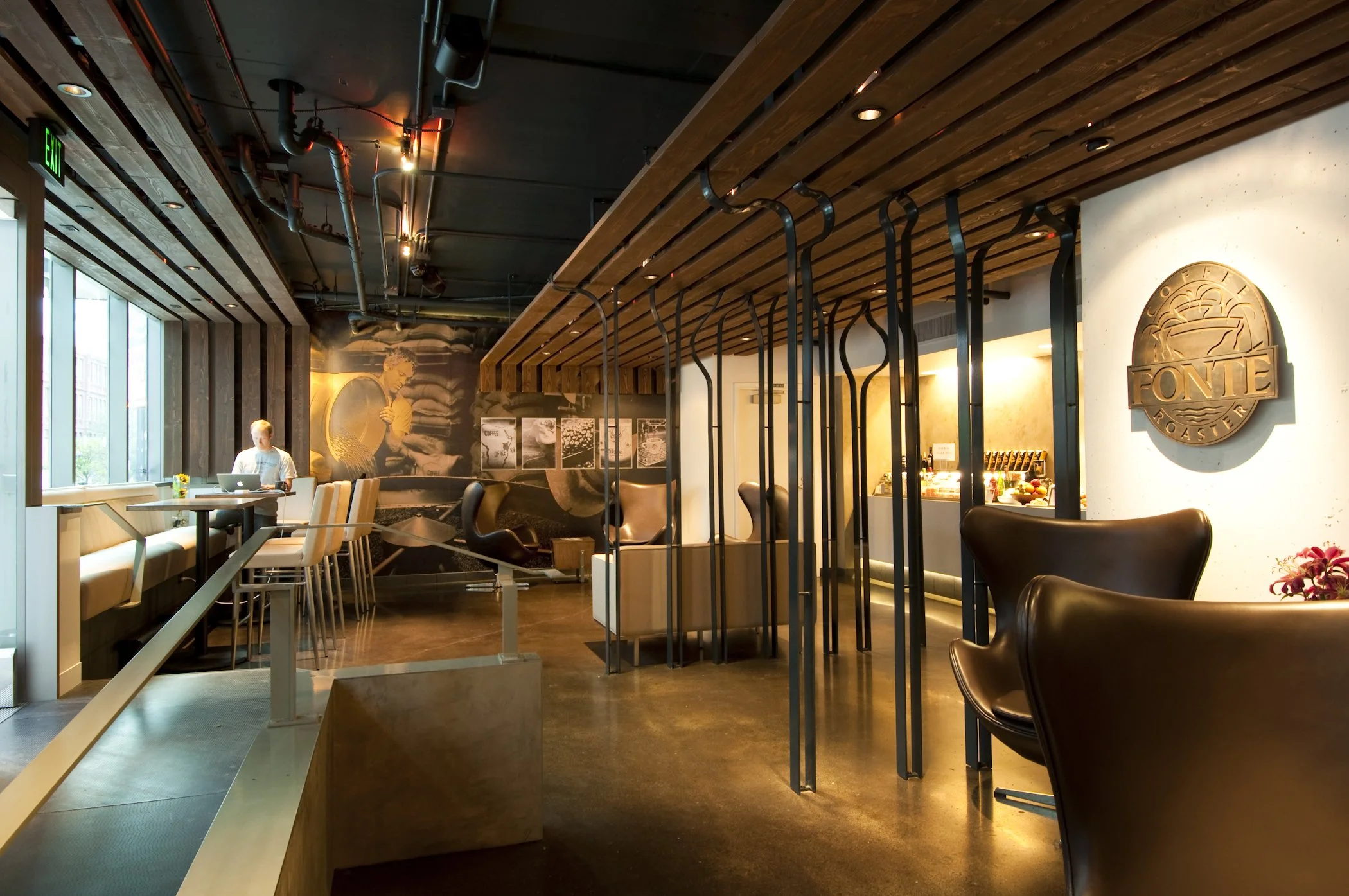 Modern coffee shop interior with wooden ceiling, leather chairs, and large wall mural. A person is seated with a laptop at a high table near a window. Fonte Coffee Roaster logo on the wall.