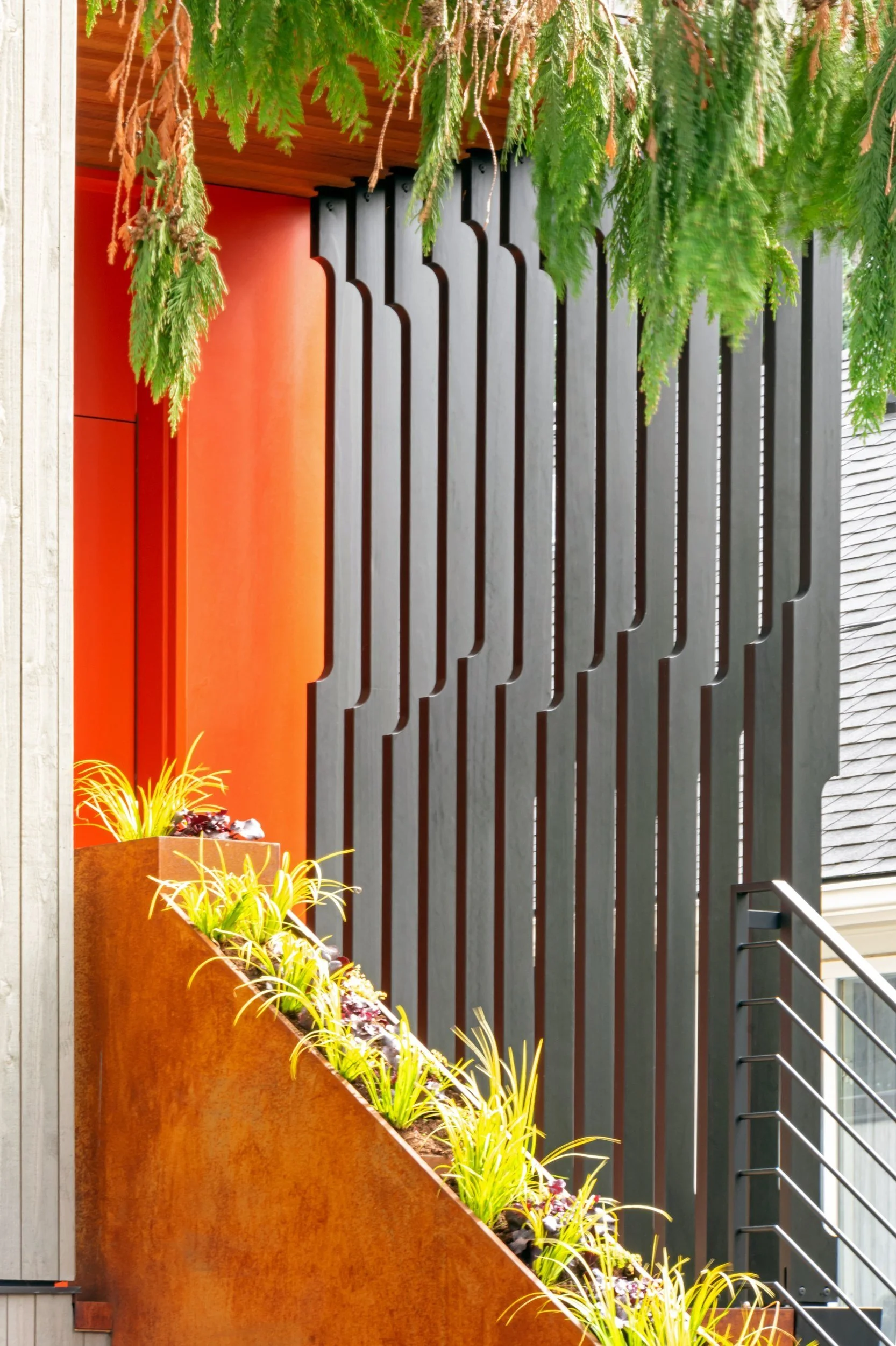 Vertical black slats in front of a vibrant orange wall, with a planter box containing green and purple plants along a rust-colored staircase. Overhanging branches add greenery to the composition.
