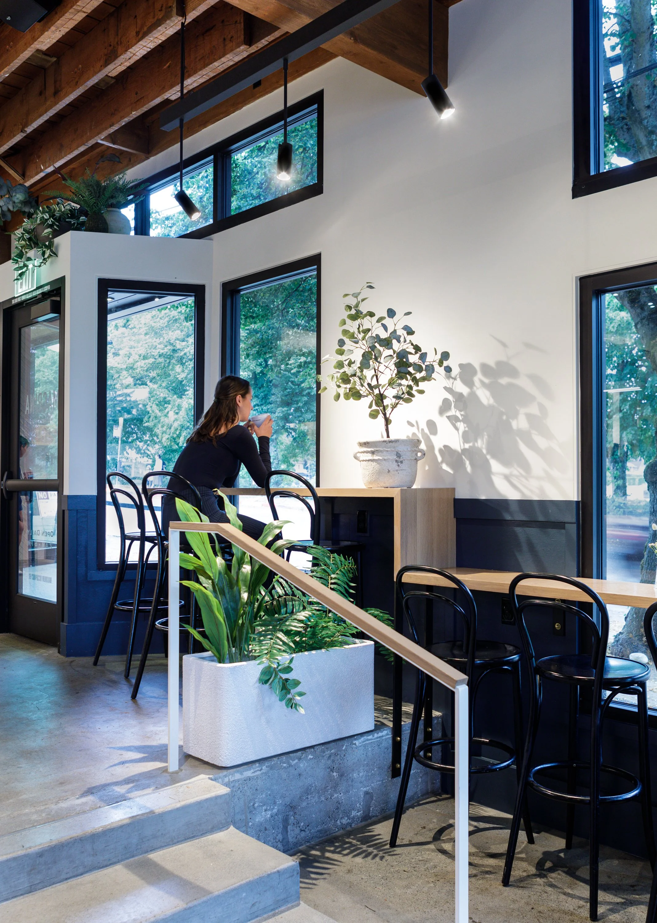 Interior of a modern cafe with a woman sitting at a counter near large windows, drinking from a cup. The space features wooden beams, potted plants, black chairs, and a minimalist design.