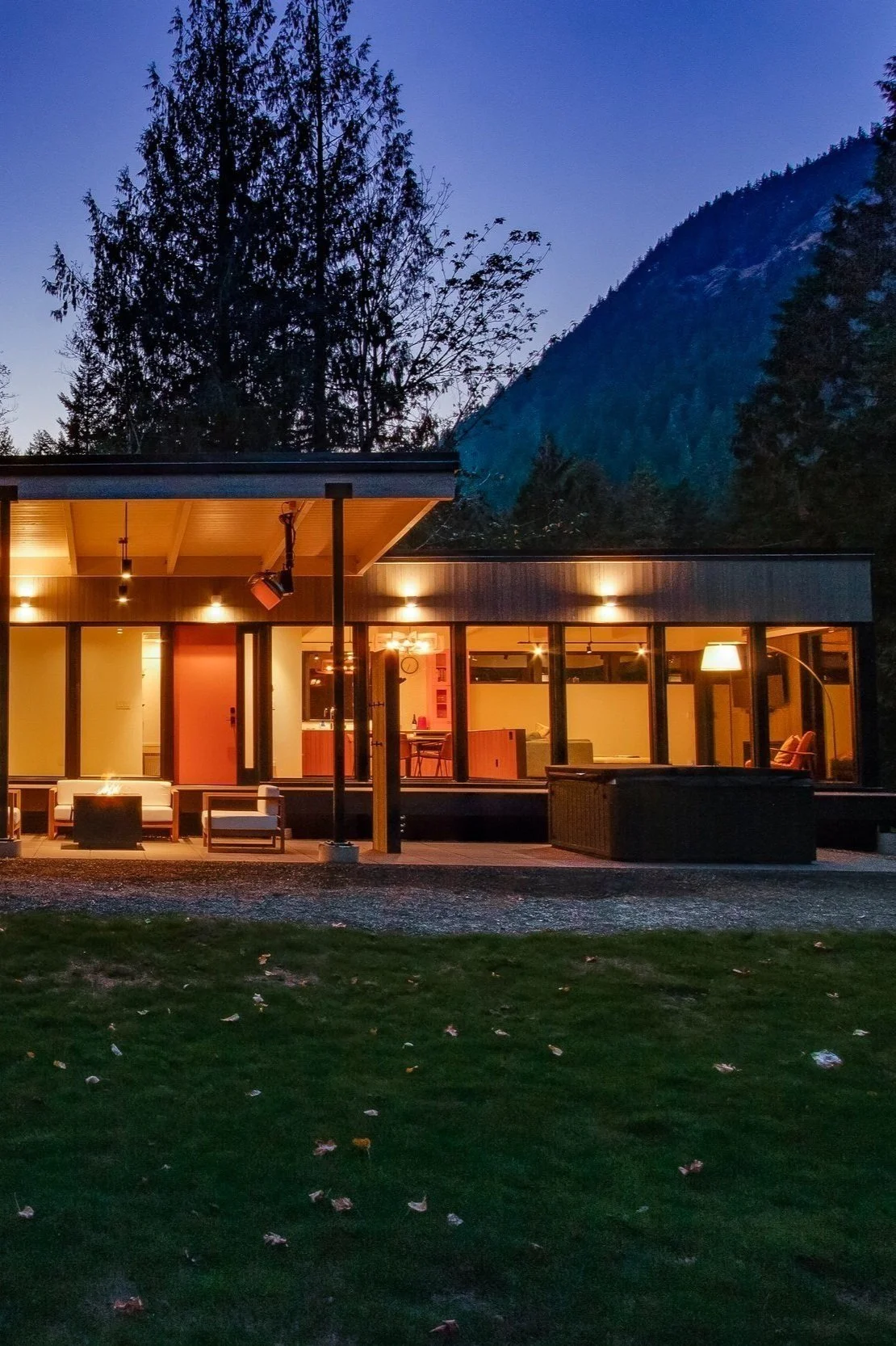 Modern house with large windows lit up at dusk, surrounded by trees and mountains.