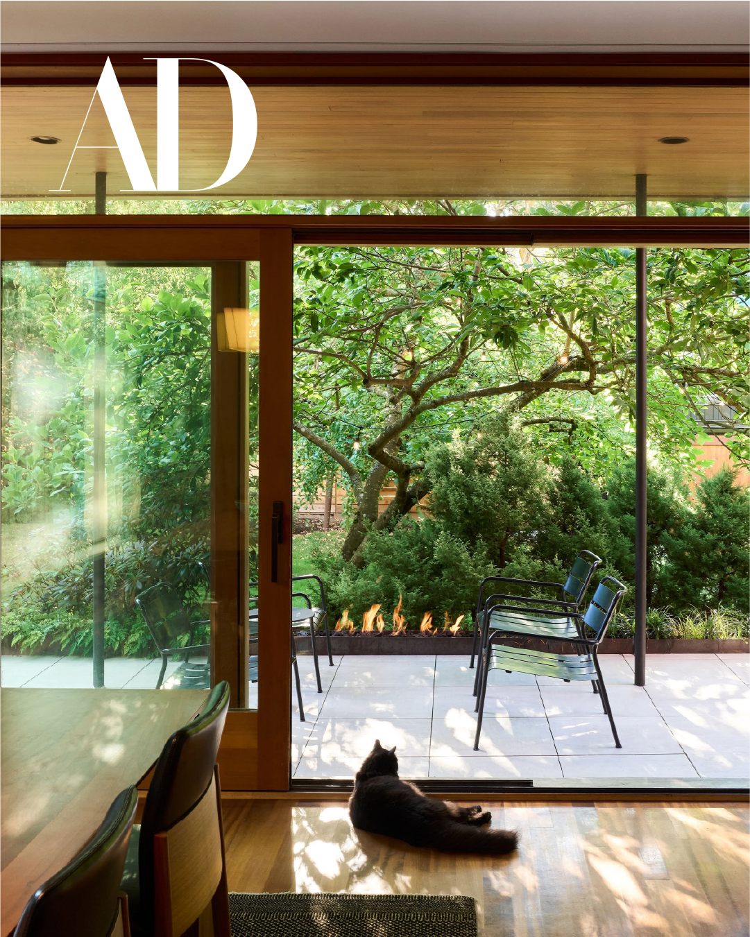 Dining room with sliding glass door opening to an outdoor patio with a custom steel fireplace and planter. Featured in Architectural Digest. A black cat is lying on the wooden floor near the door, watching the garden.
