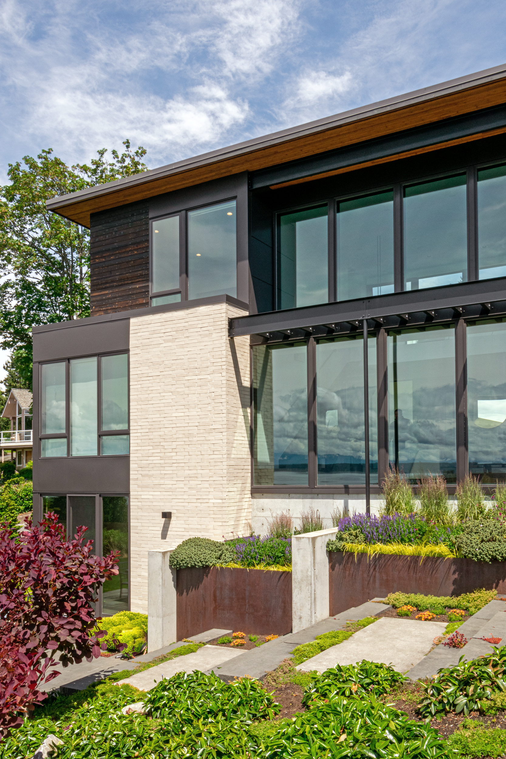 Modern house with large windows, stone and wood siding, surrounded by landscaped garden with various plants, under a partly cloudy sky.