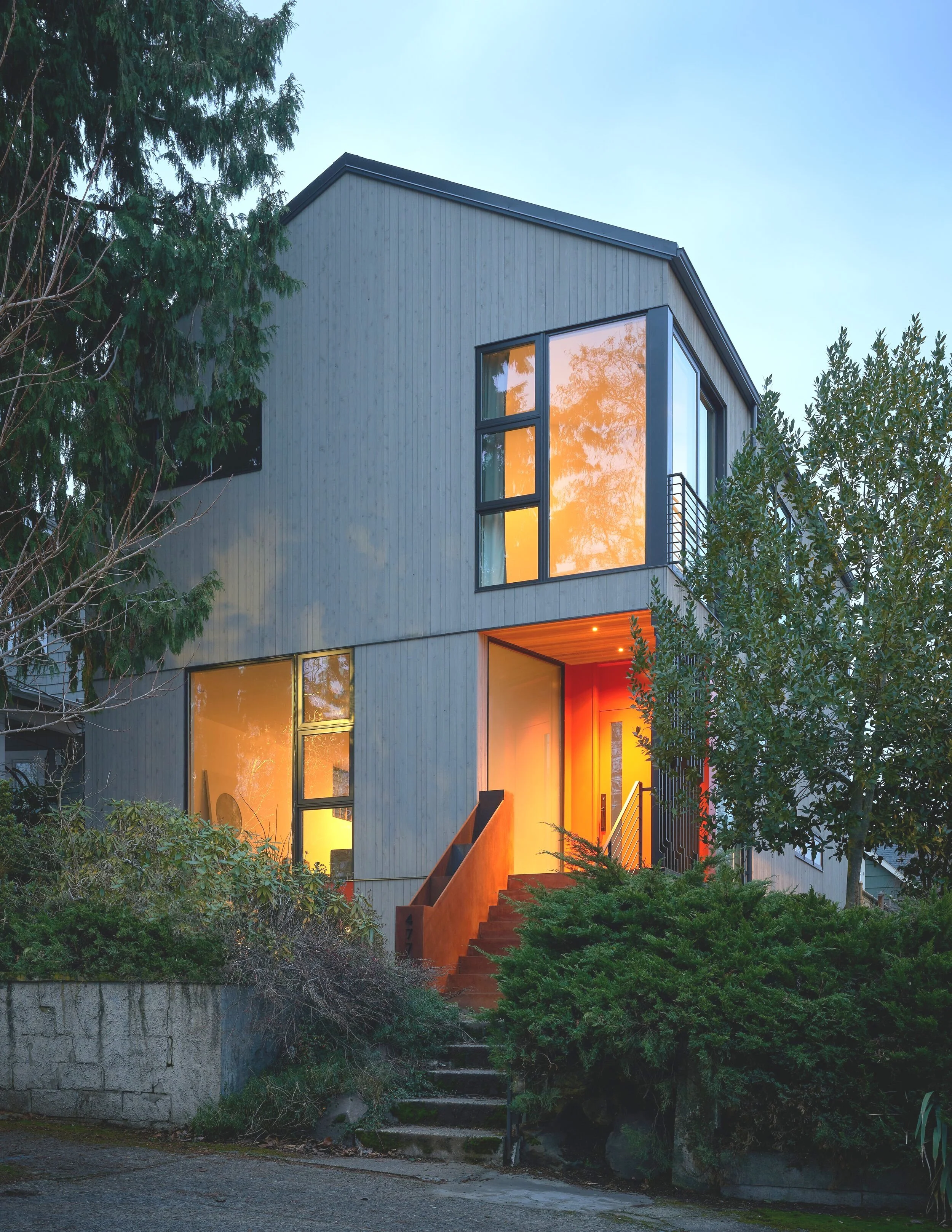 Modern two-story house with large windows and illuminated interior, surrounded by trees and shrubs.