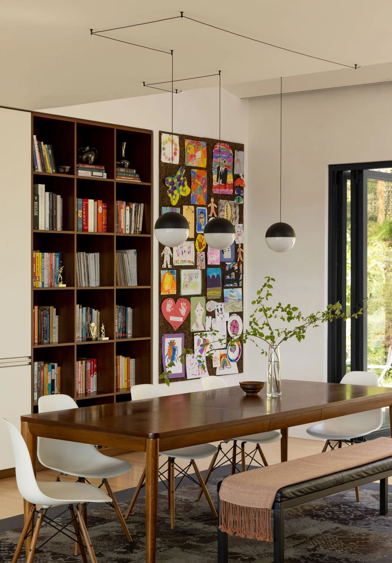 Modern dining room with wooden table, white chairs, pendant lights, a wall of children’s artwork, and bookshelf with books and decor items.