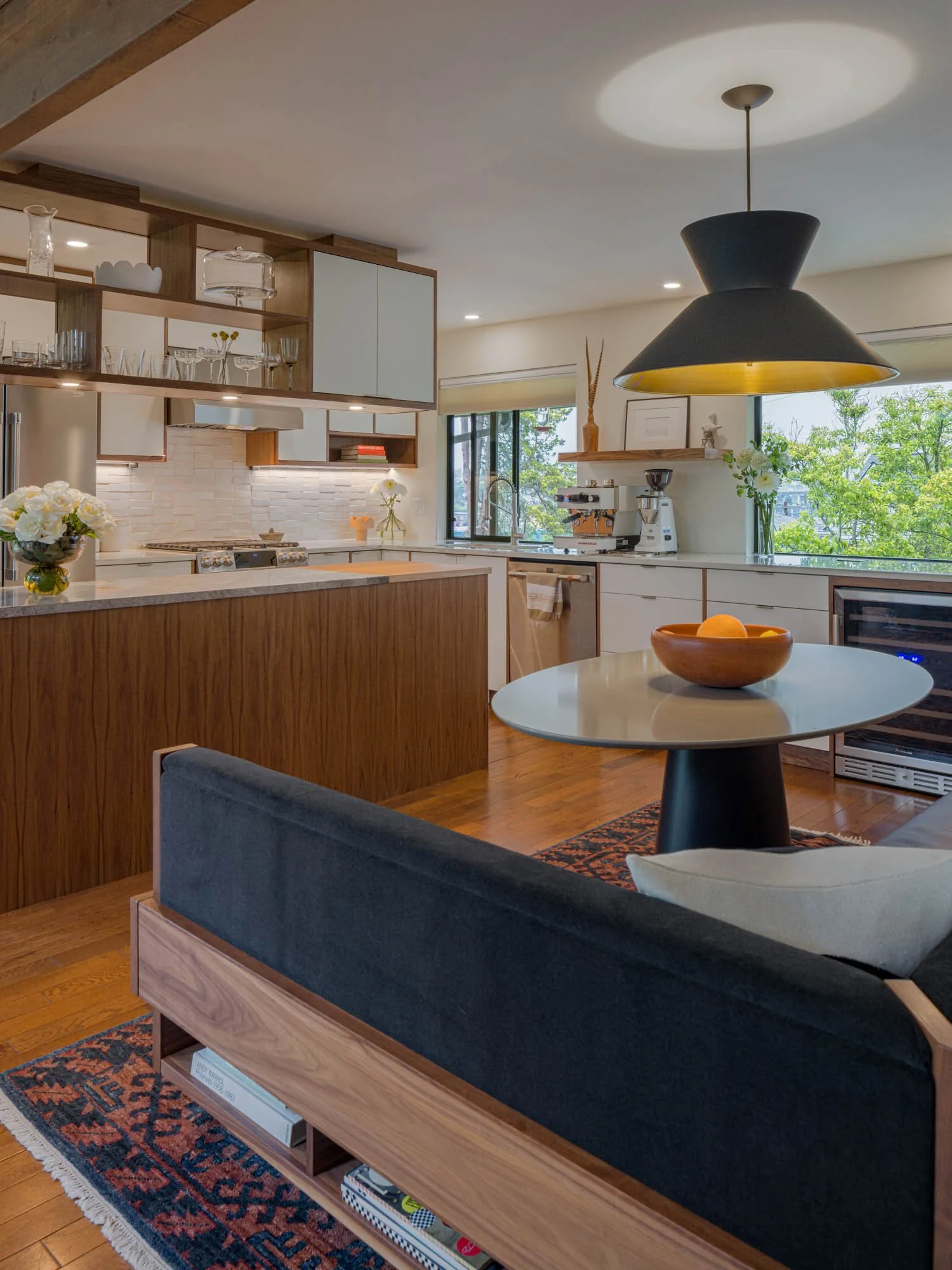 Modern kitchen with wooden cabinets, white countertops, and a large black pendant light. Includes open shelving with glassware, a coffee maker near the window, and a small dining area with a table and bowl of oranges. A dark couch and patterned rug complete the space.