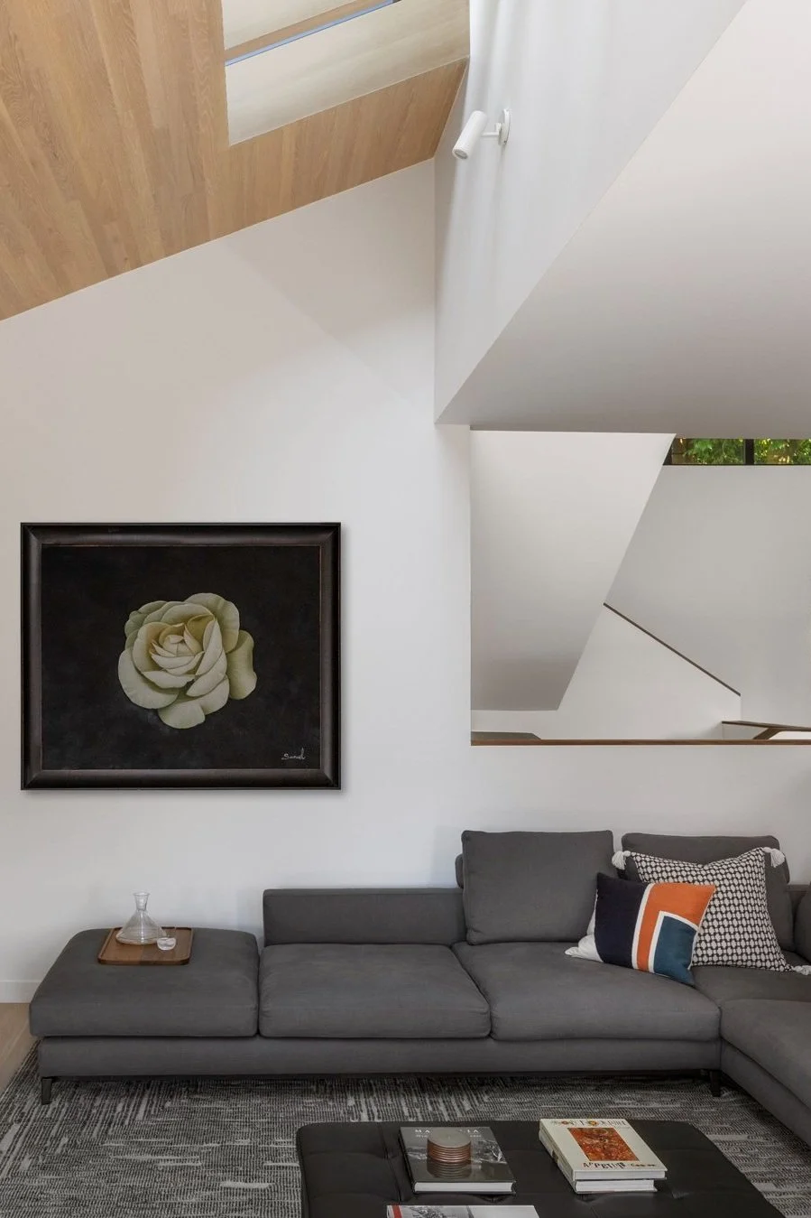 Modern living room with a gray sectional sofa, geometric throw pillows, and a large floral painting on the wall. The space features wooden accents, minimalist design, and a small coffee table with books and a decorative item.