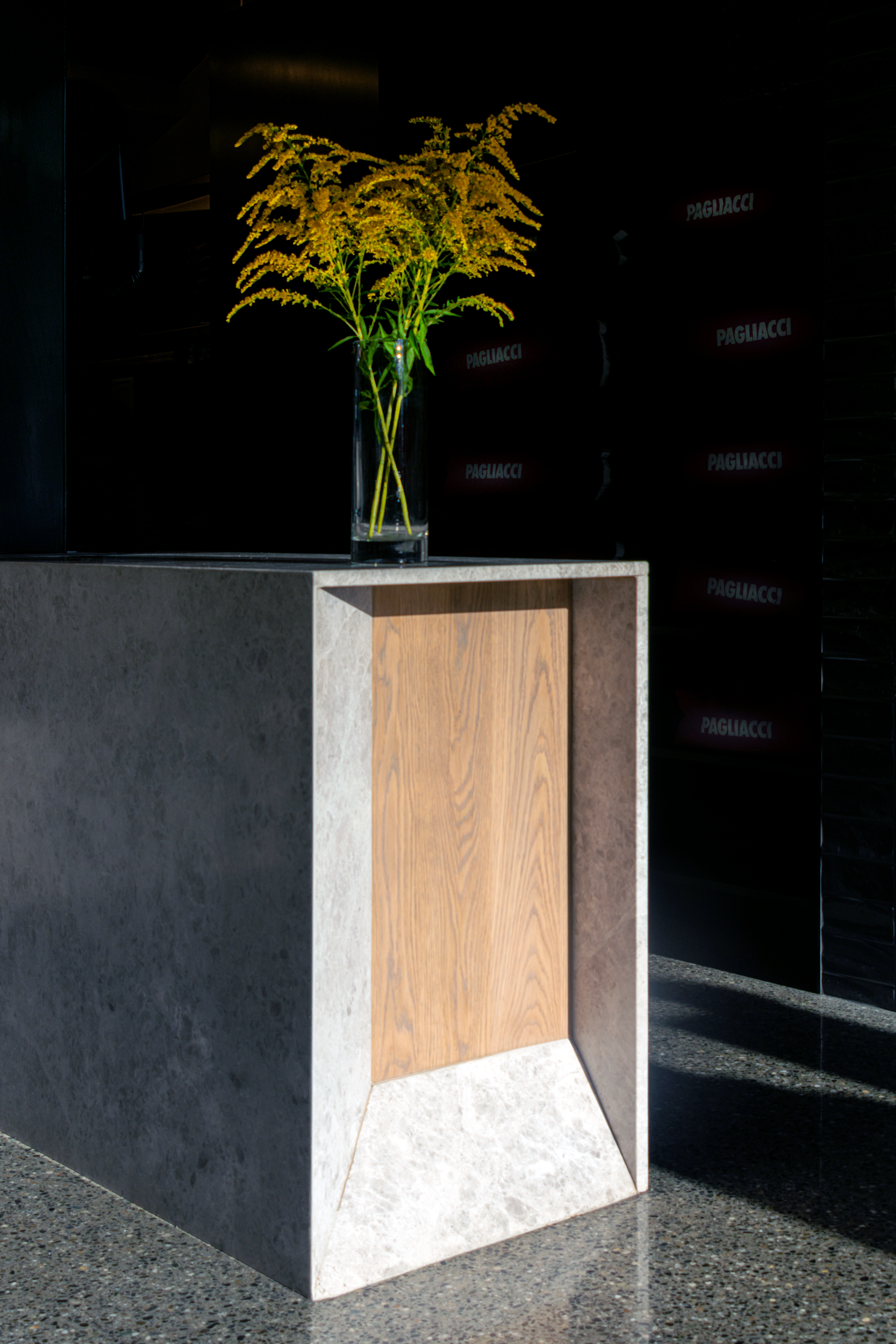 Vase with yellow flowers on a modern concrete and wooden pedestal, dark background with subtle signage.