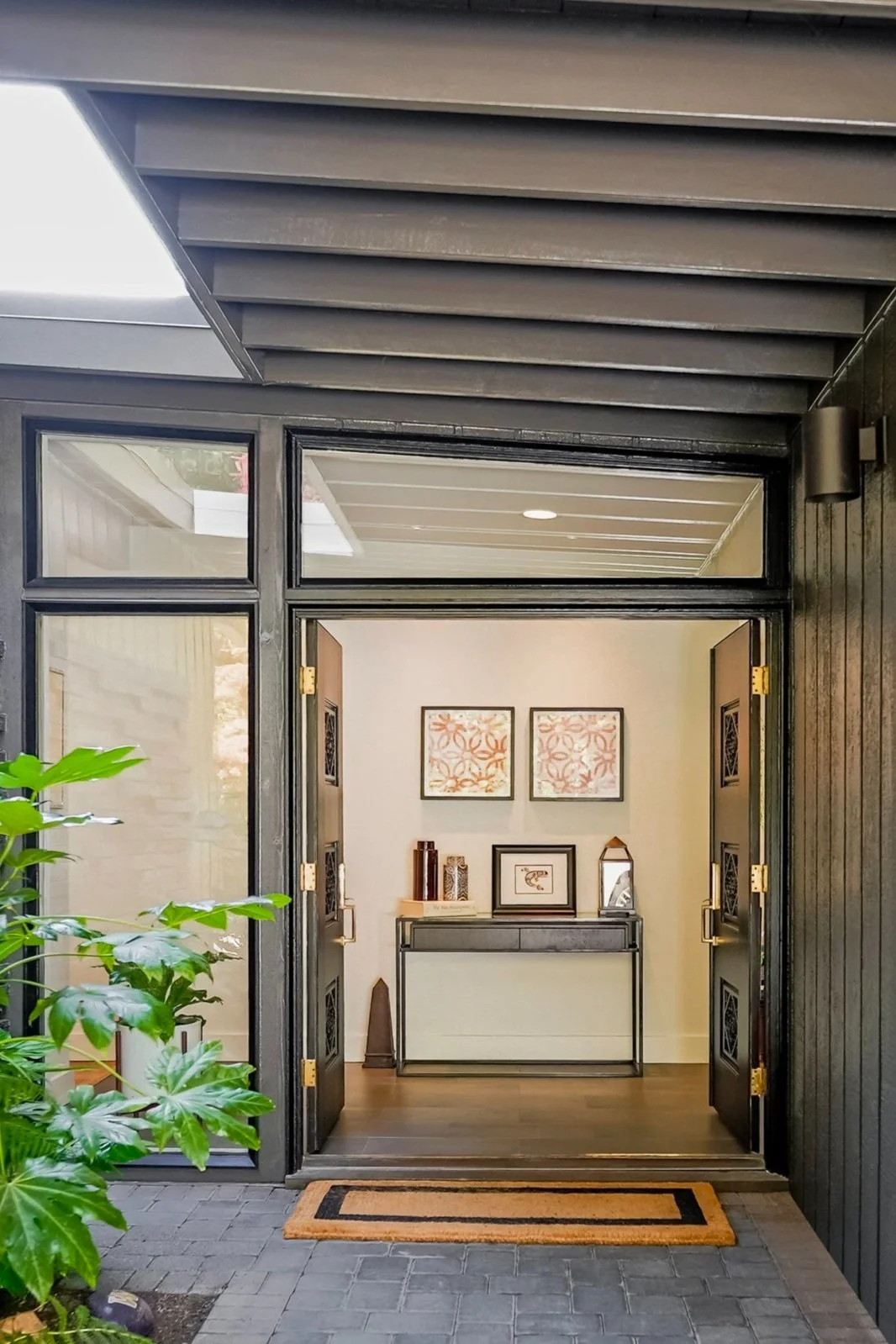 Open front door of a modern house with black framing, leading to an entrance hallway. Interior decor includes wall art, a console table with decorative items, and indoor plants.