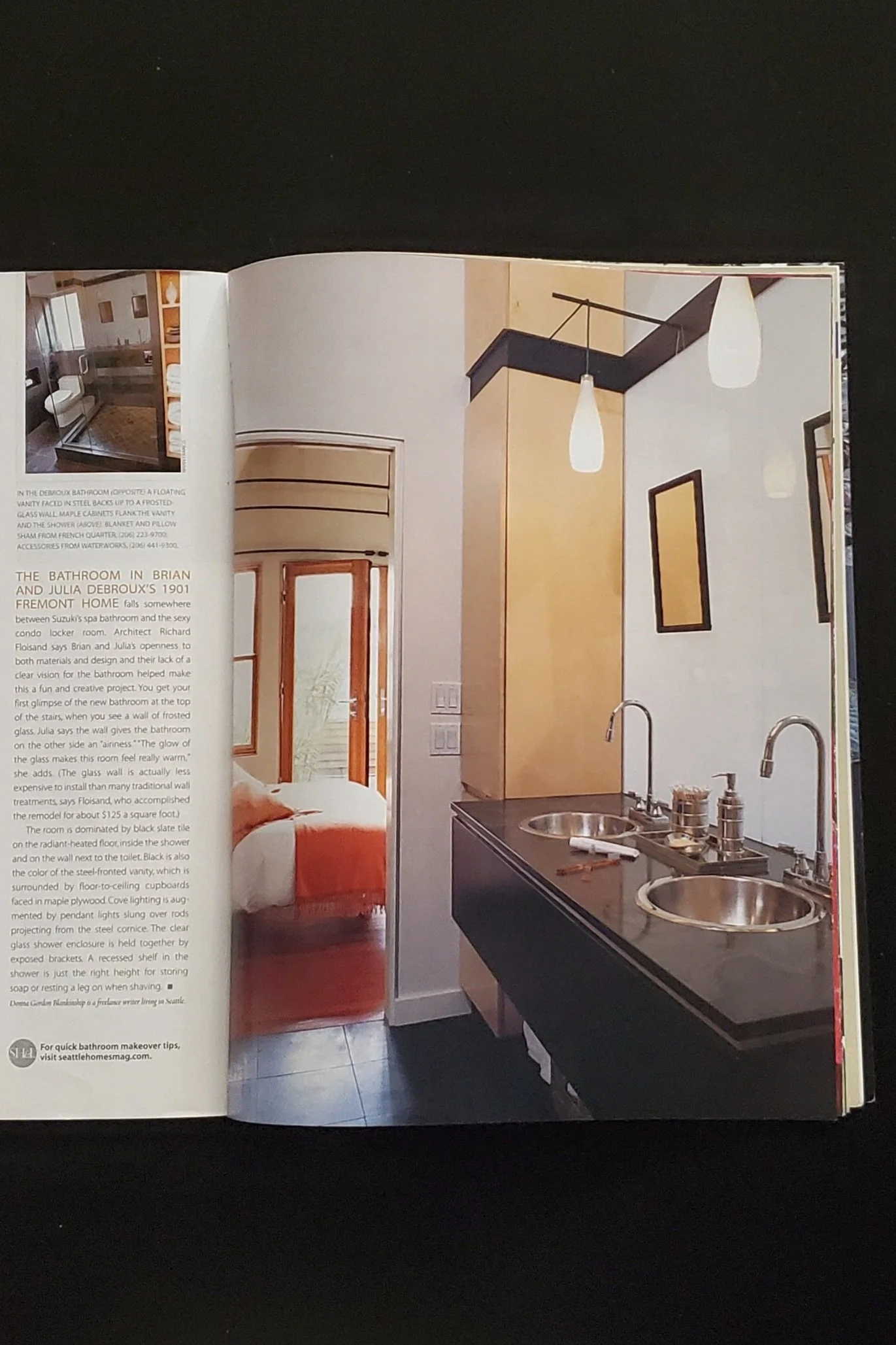 Open magazine showing a modern bathroom with dual sinks, pendant lighting, and an adjacent bedroom. The layout features sleek cabinetry and black countertops, with a small picture insert of another bathroom space on the left page.