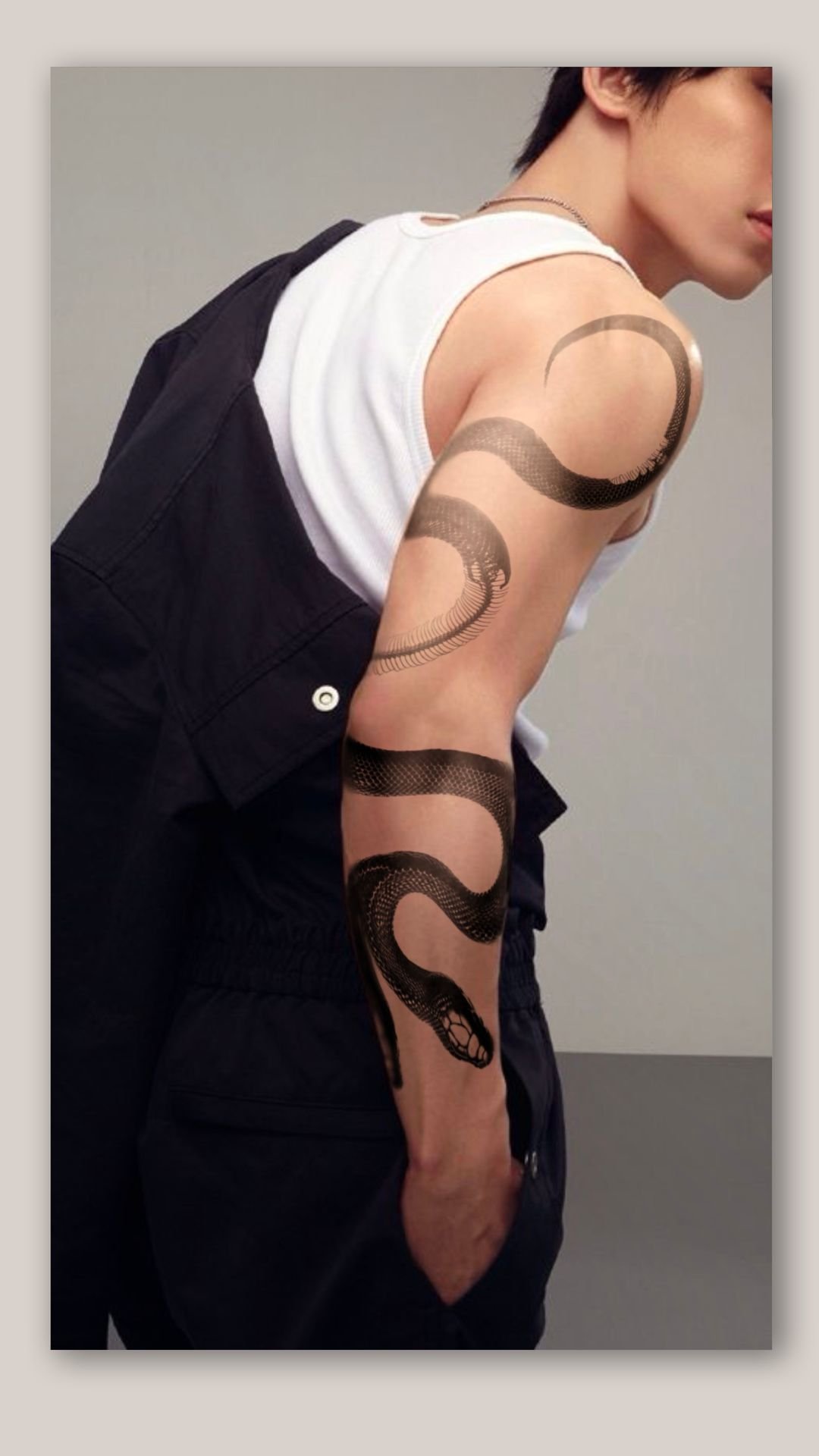 A person with short black hair and a snake tattoo on their arm, wearing a white sleeveless shirt and black pants, leaning forward with their hand in their pocket, against a plain gray background.