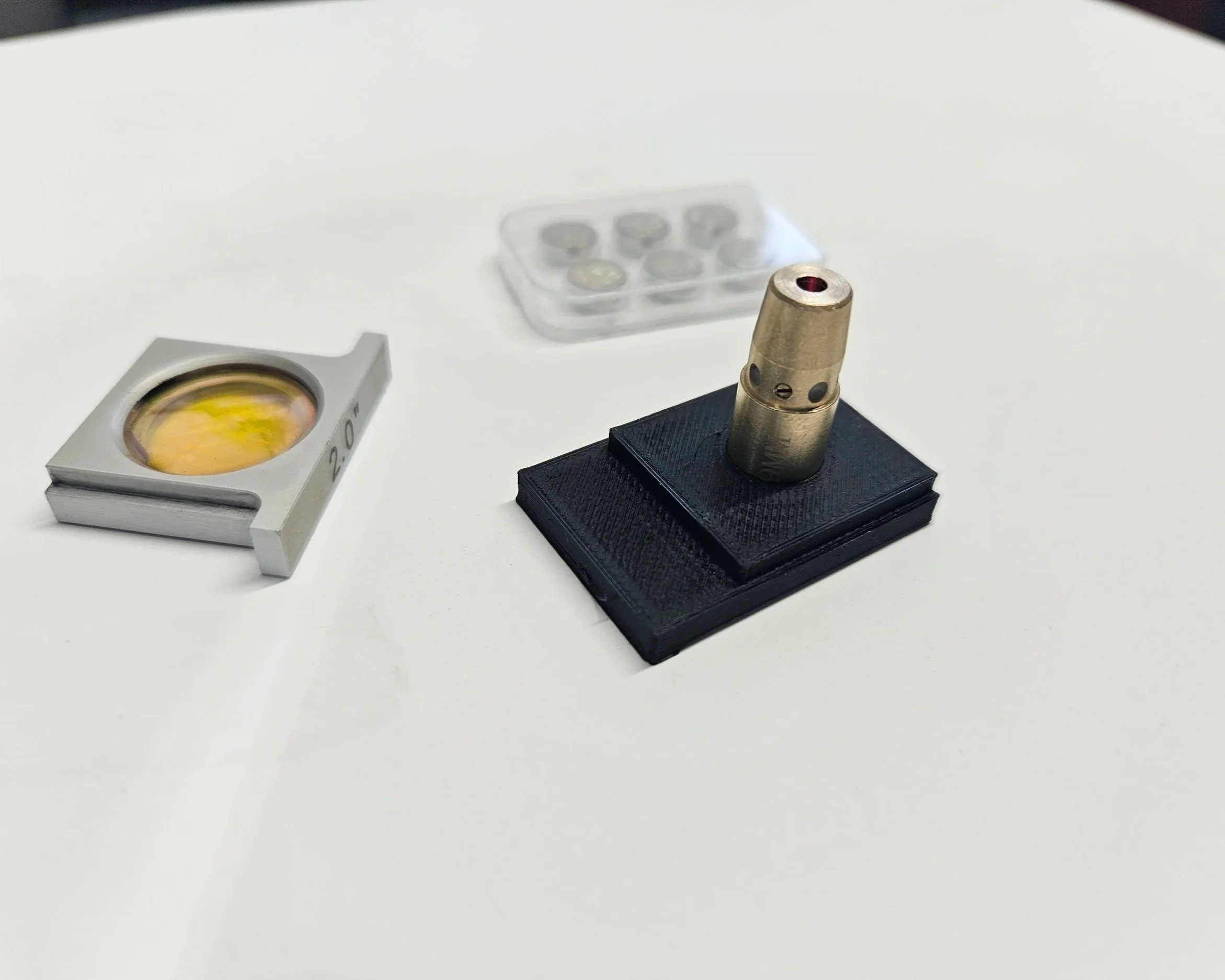 A small metal device with a red dot on top, mounted on a black rectangular base, with a clear plastic container of small items and another metallic object with a transparent window nearby, all on a white surface.