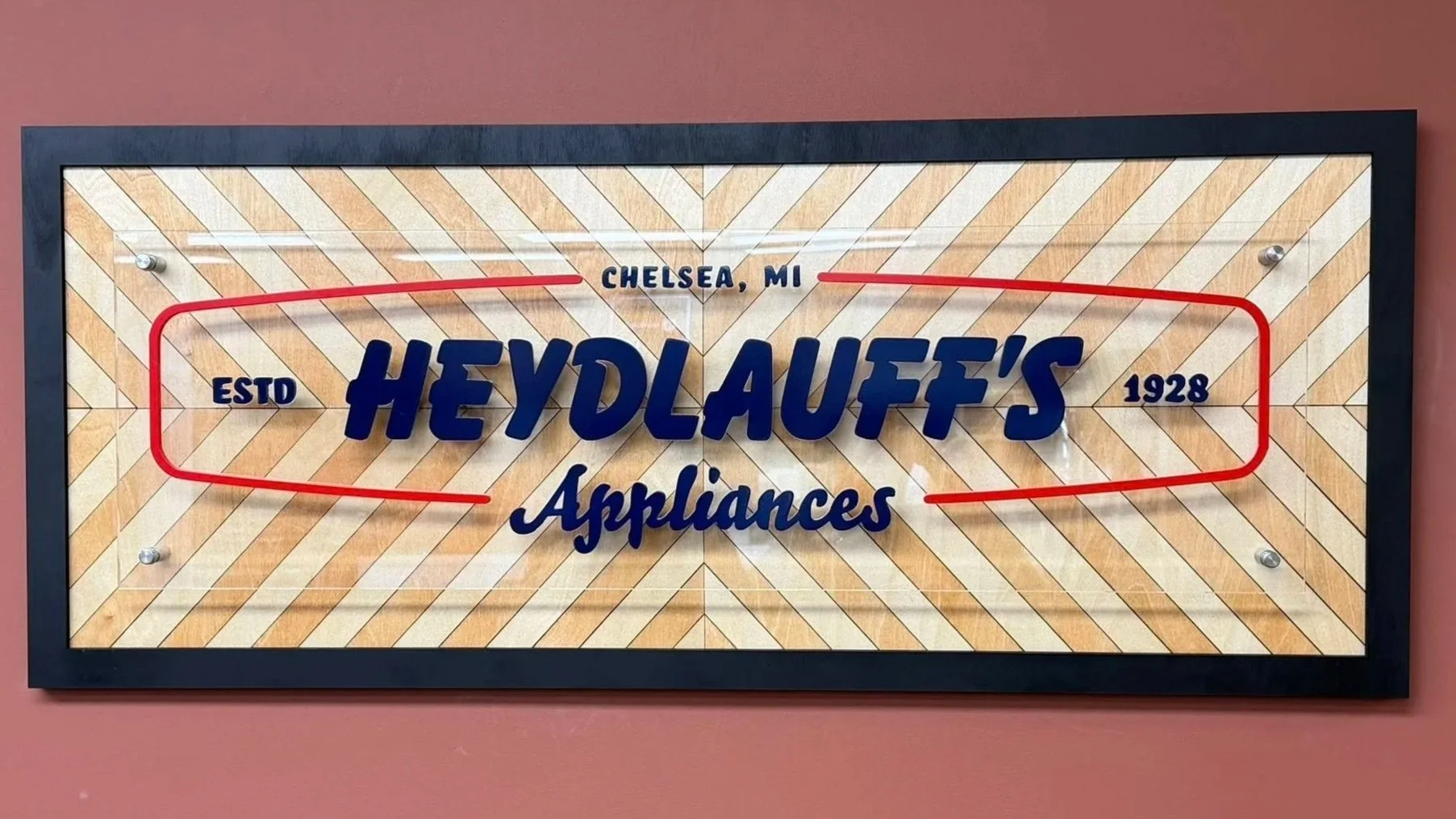 Framed sign with vintage design reading "Heydlauff's Appliances, Estd 1928, Chelsea, MI" on a pink wall.